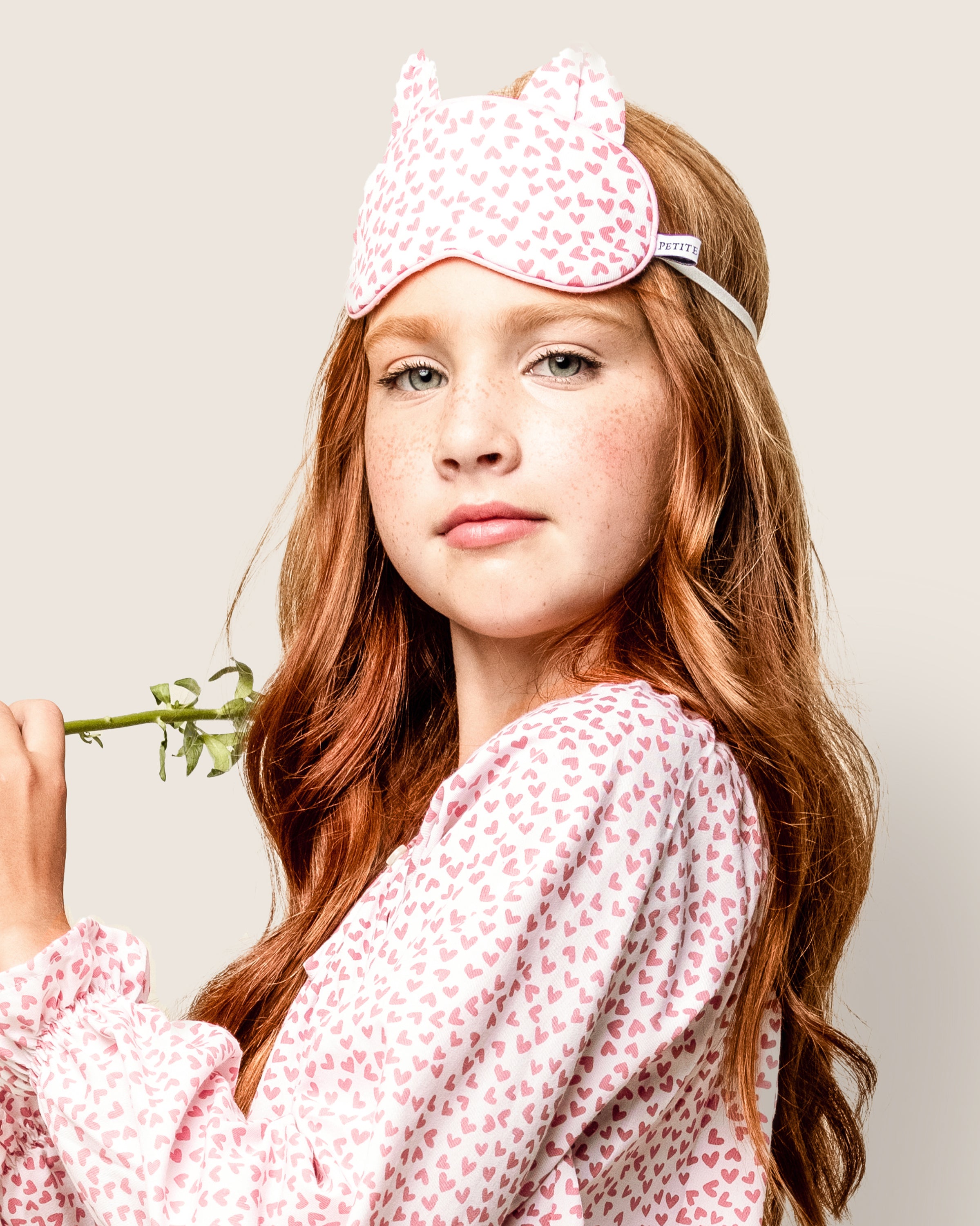 A young girl with long red hair wears Petite Plumes Kids Kitty Sleep Mask in Sweethearts and a pajama top with heart patterns. She holds a small flower stem and gazes at the camera neutrally.