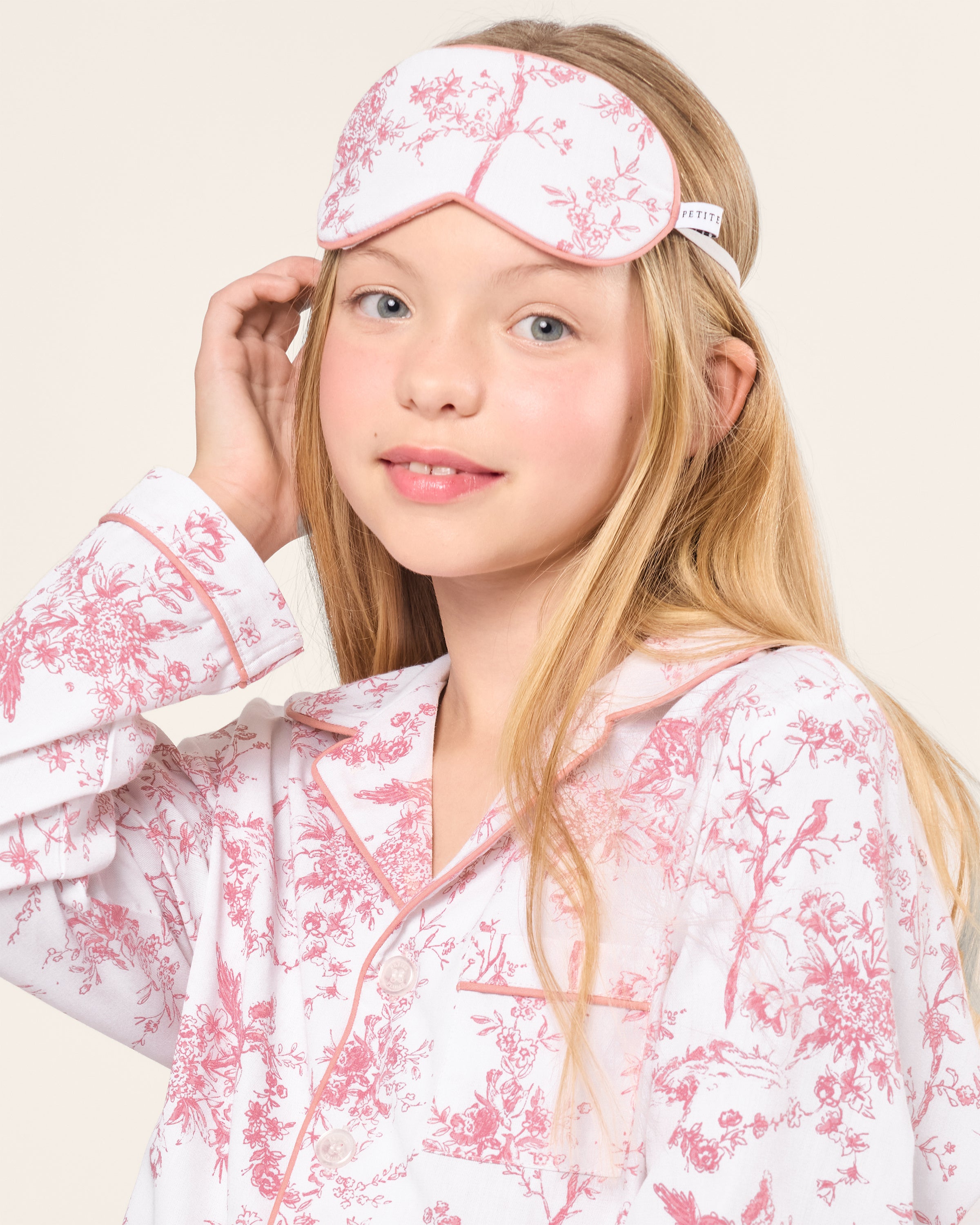 A young girl with long blonde hair wears the Kids Twill Sleep Mask in Pink Timeless Toile by Petite Plume, paired with a matching white and pink patterned pajama set adorned with botanical flowers. She smiles softly, touching the side of her head against a plain, light-colored background.