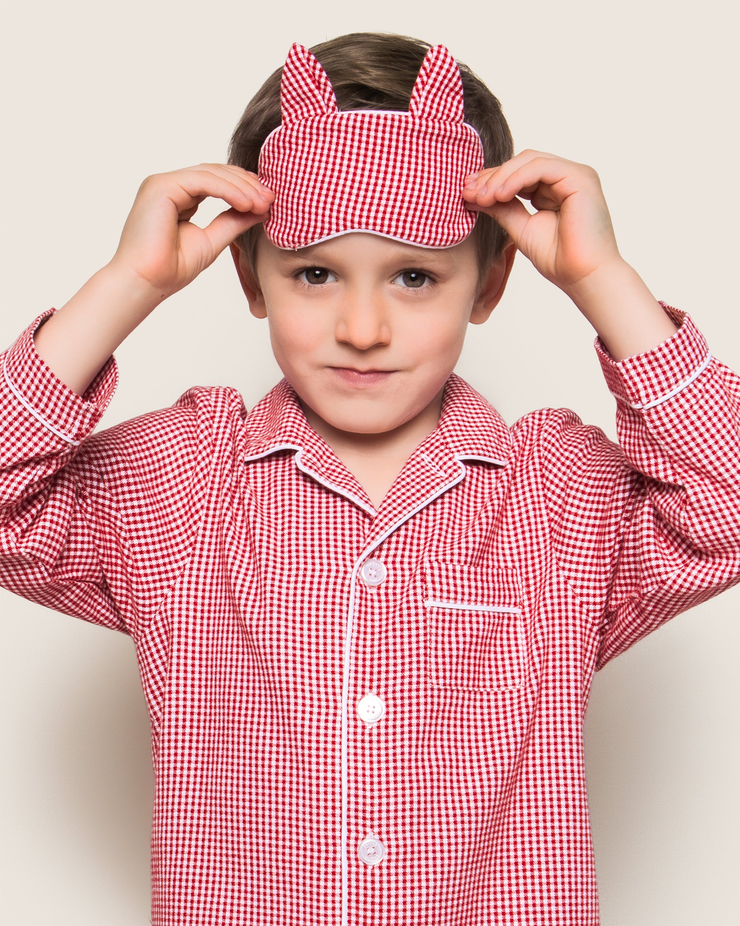 In a serene moment against a plain backdrop, a child adjusts the Petite Plume Kids Flannel Red Mini-Gingham Kitty Sleep Mask atop their red checkered pajamas, which feature piping along the collar, button placket, and a chest pocket.