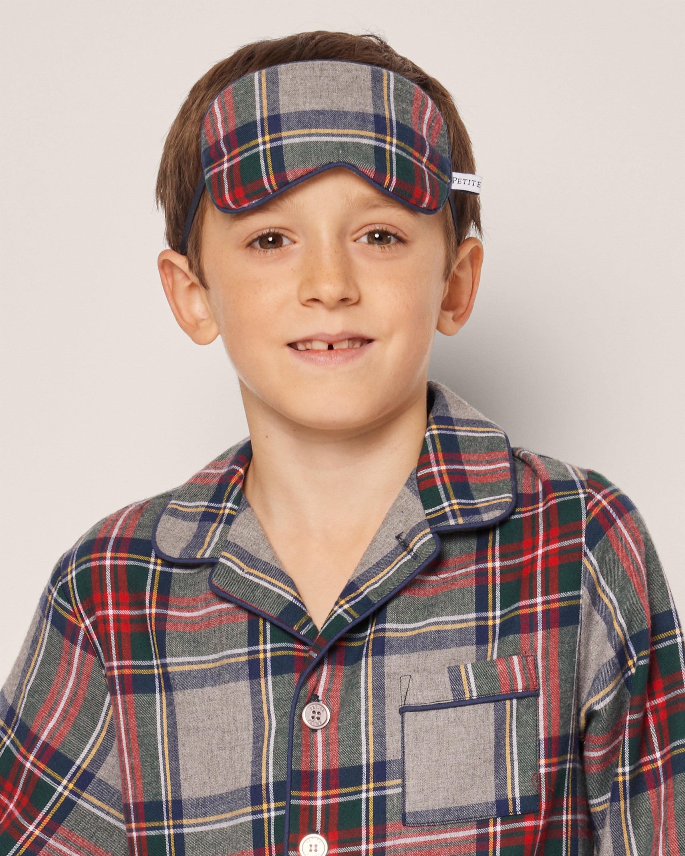 A young boy in plaid pajamas and Petite Plumes Kids Brushed Cotton Sleep Mask in Westminster Tartan, featuring a colorful red, blue, and gray design, looks at the camera against a plain white background.