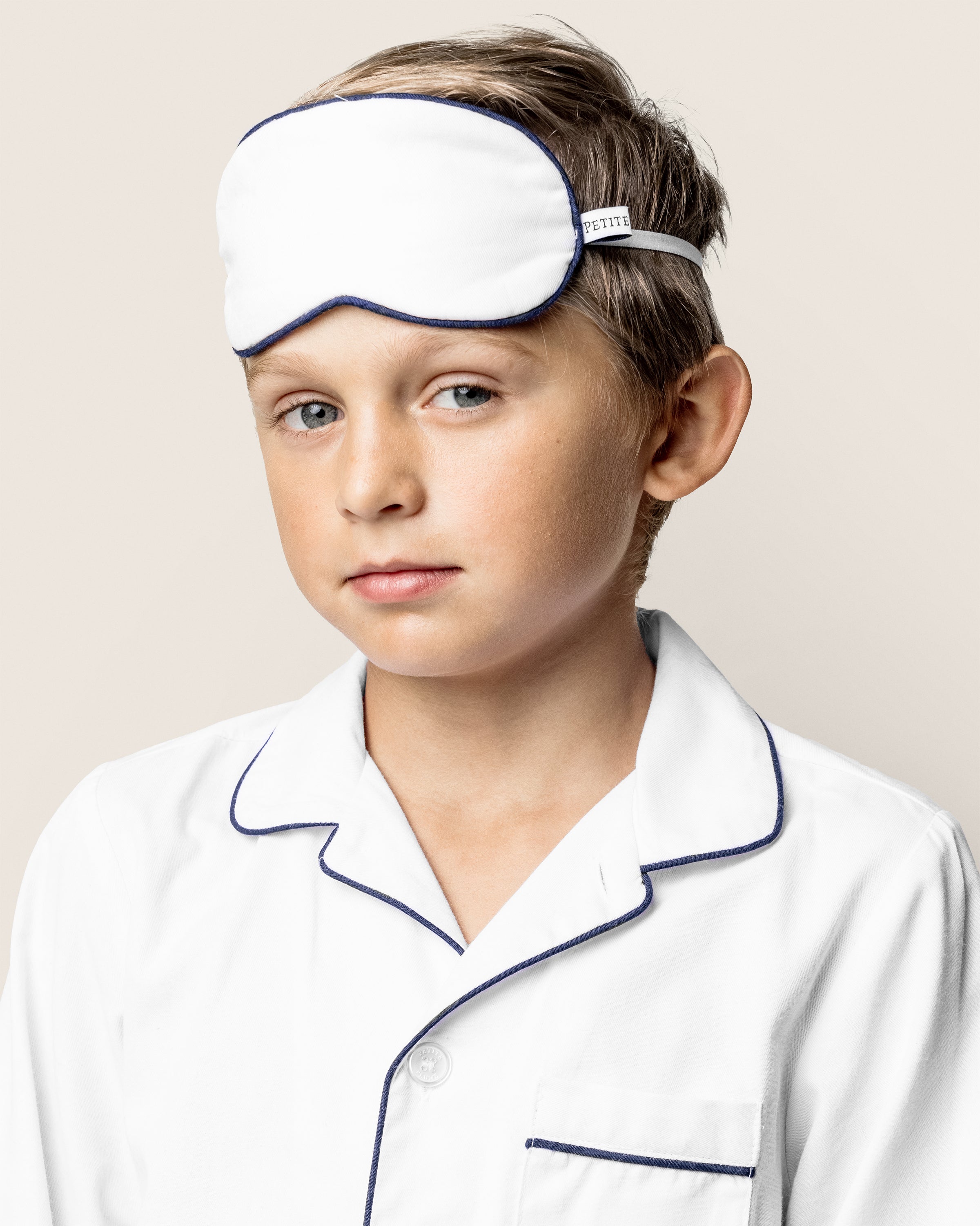 A child, in Petite Plumes Kids Twill Sleep Mask in White with Navy Piping, stands against a plain background. Their short hair complements the relaxed expression that radiates comfort and calm.