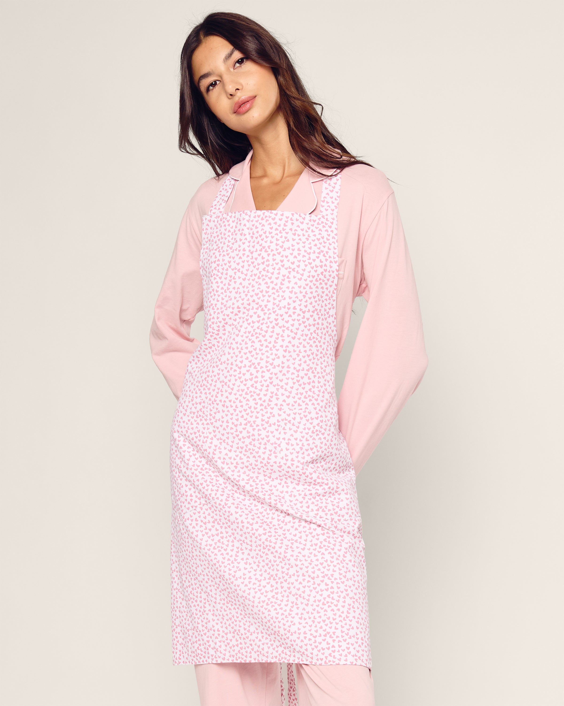 Wearing Petite Plumes Adult Apron in Sweethearts over a light pink long-sleeve shirt and pants, a woman stands confidently with hands on her hips against a plain background, capturing a modern timeless style.