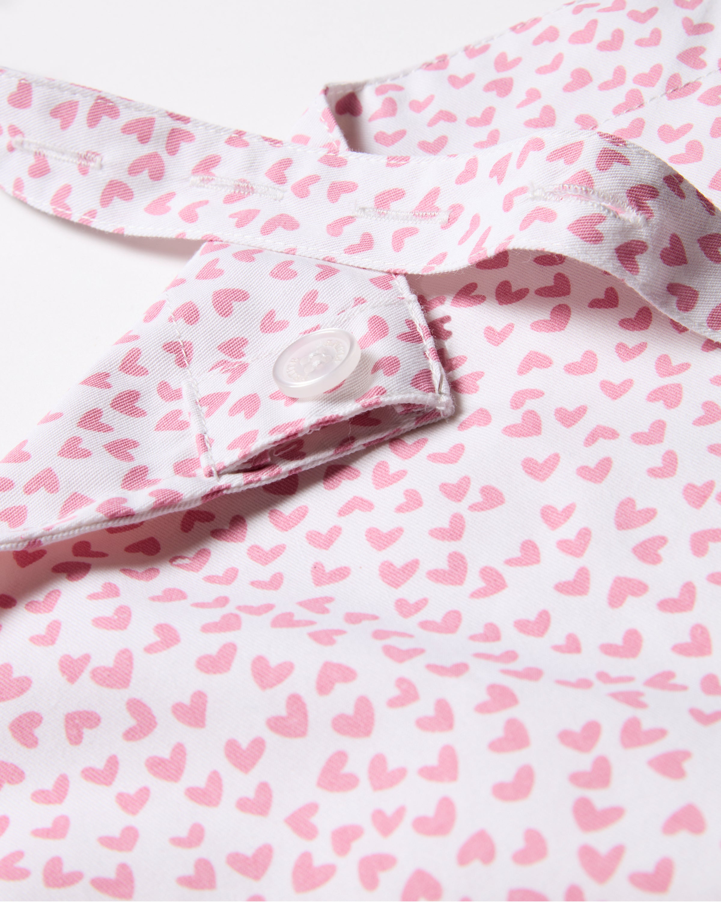 The image features the Adult Apron in Sweethearts by Petite Plume, highlighting its modern timeless aesthetic with a white fabric adorned in pink heart patterns and a buttoned strap, showcasing its design and texture.