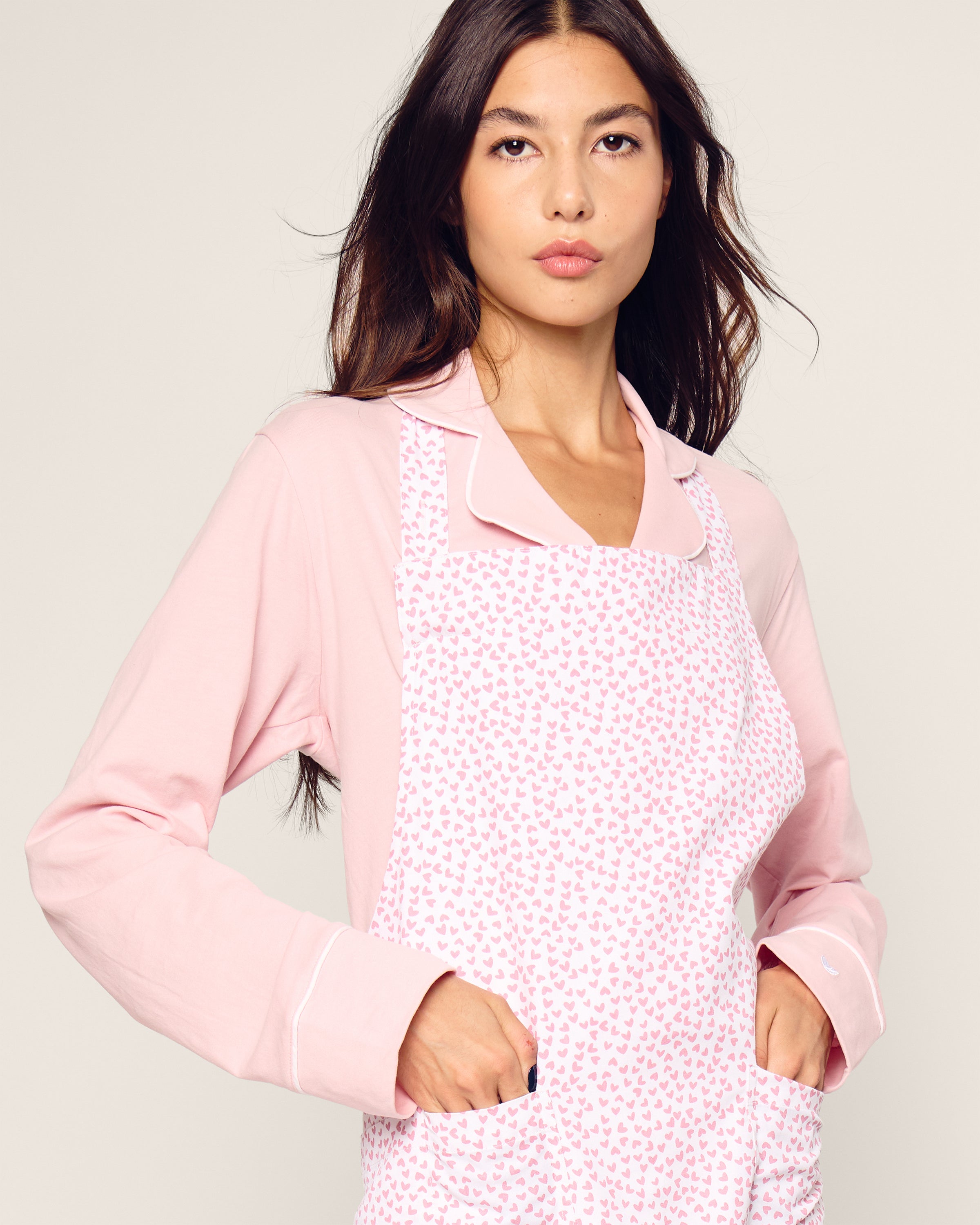 A person with long dark hair wears a modern, timeless pink collared shirt and the Petite Plume Adult Apron in Sweethearts. With hands resting in the apron pockets, they gaze forward with a neutral expression.