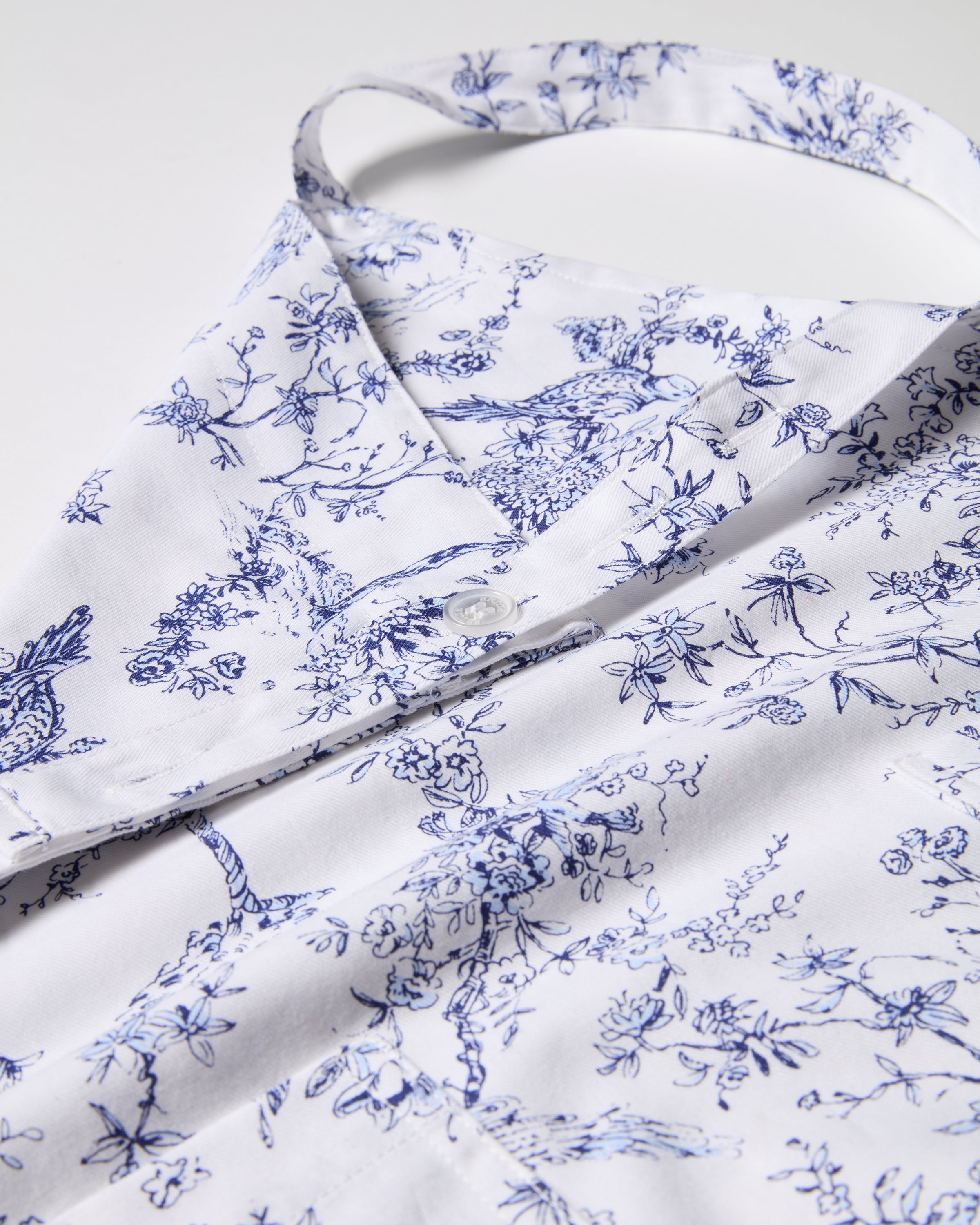 Close-up of a white Adult Apron in Timeless Toile by Petite Plume, featuring an intricate blue floral design. Detailed flower and branch drawings create delicate beauty. Laid flat, it reveals buttons and a neck loop, ideal for holiday baking.