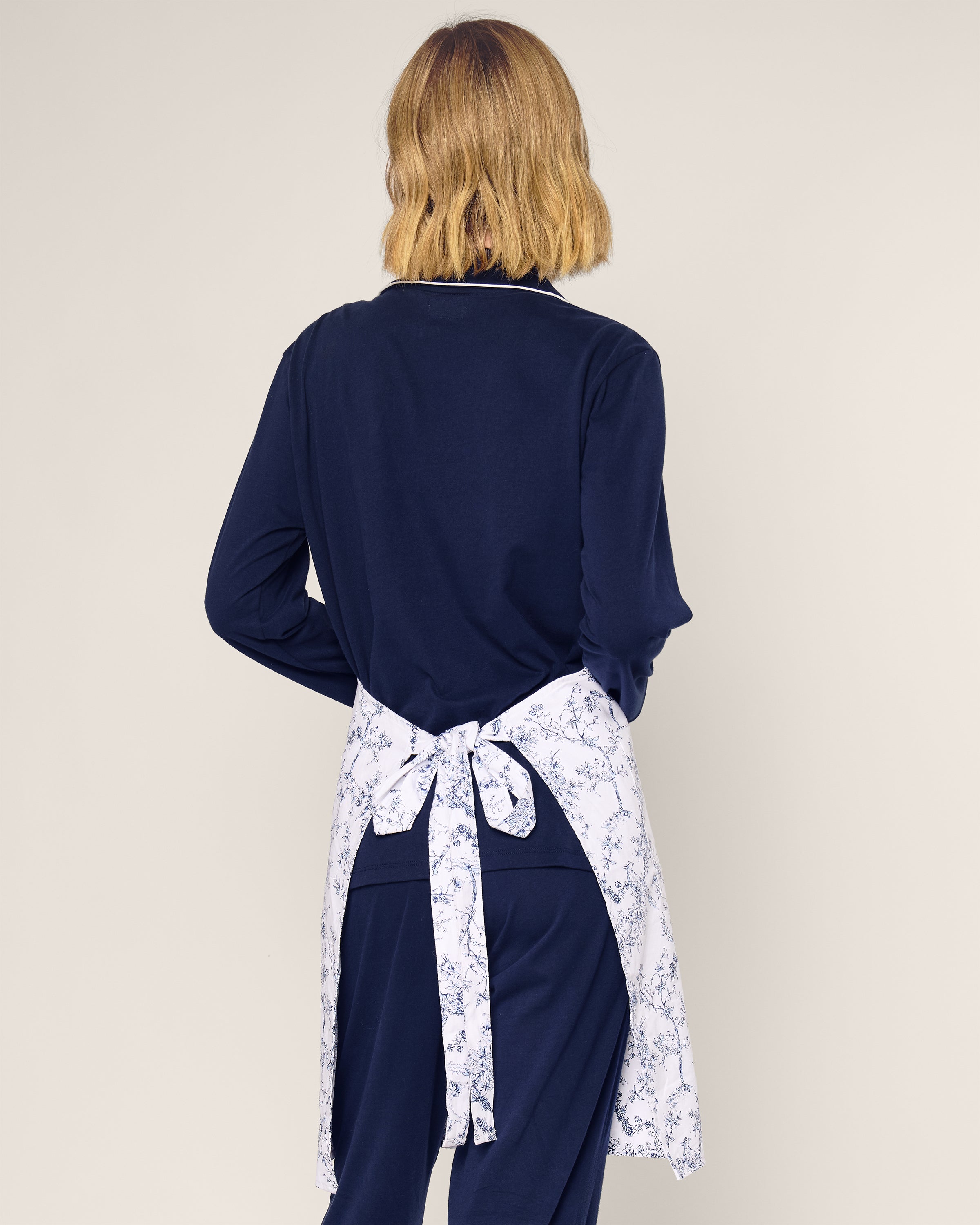 A person with shoulder-length blond hair, seen from behind in a navy outfit, wears a Petite Plume Adult Apron in Timeless Toile, tied at the back and exuding botanical elegance. The plain background enhances the apron’s timeless beauty.