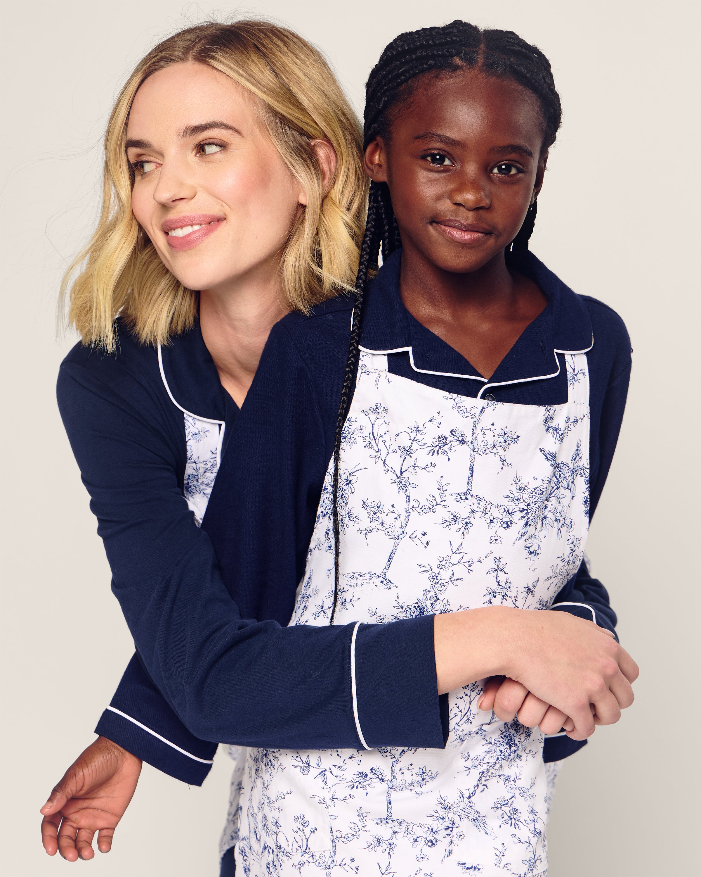 Two people wearing Timeless Toile aprons by Petite Plume, with short blonde hair on the left and long braided hair on the right, hug and look cheerfully in the same direction, exuding harmony and joy.