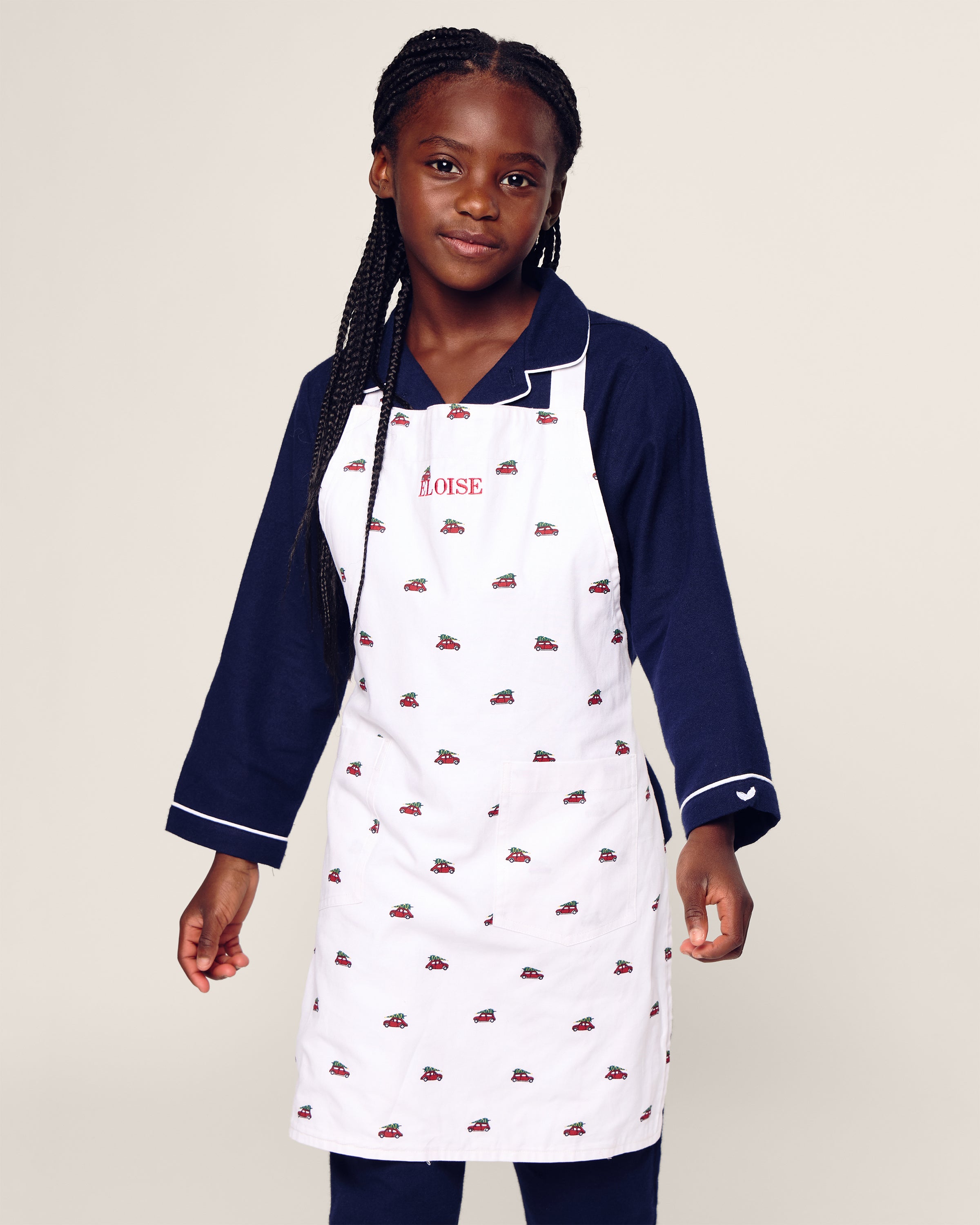 A young girl with braided hair wears Petite Plumes Kids Apron in Holiday Journey—a navy long-sleeve shirt complements the white apron with red car patterns and Eloise embroidered in red. Perfect for holiday baking, she smiles slightly against a plain background. Available in childrens sizes.