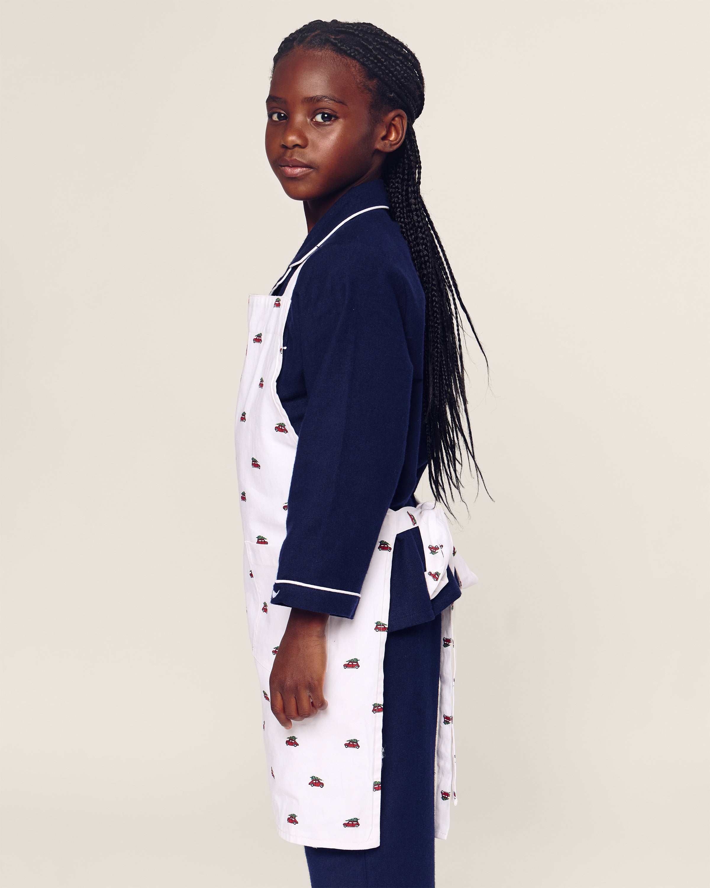 A young girl with long braided hair stands in profile, wearing a dark blue outfit and the Petite Plume Kids Apron in Holiday Journey. The white apron, featuring small red patterns, complements her look as she confidently faces the camera against a plain light background.