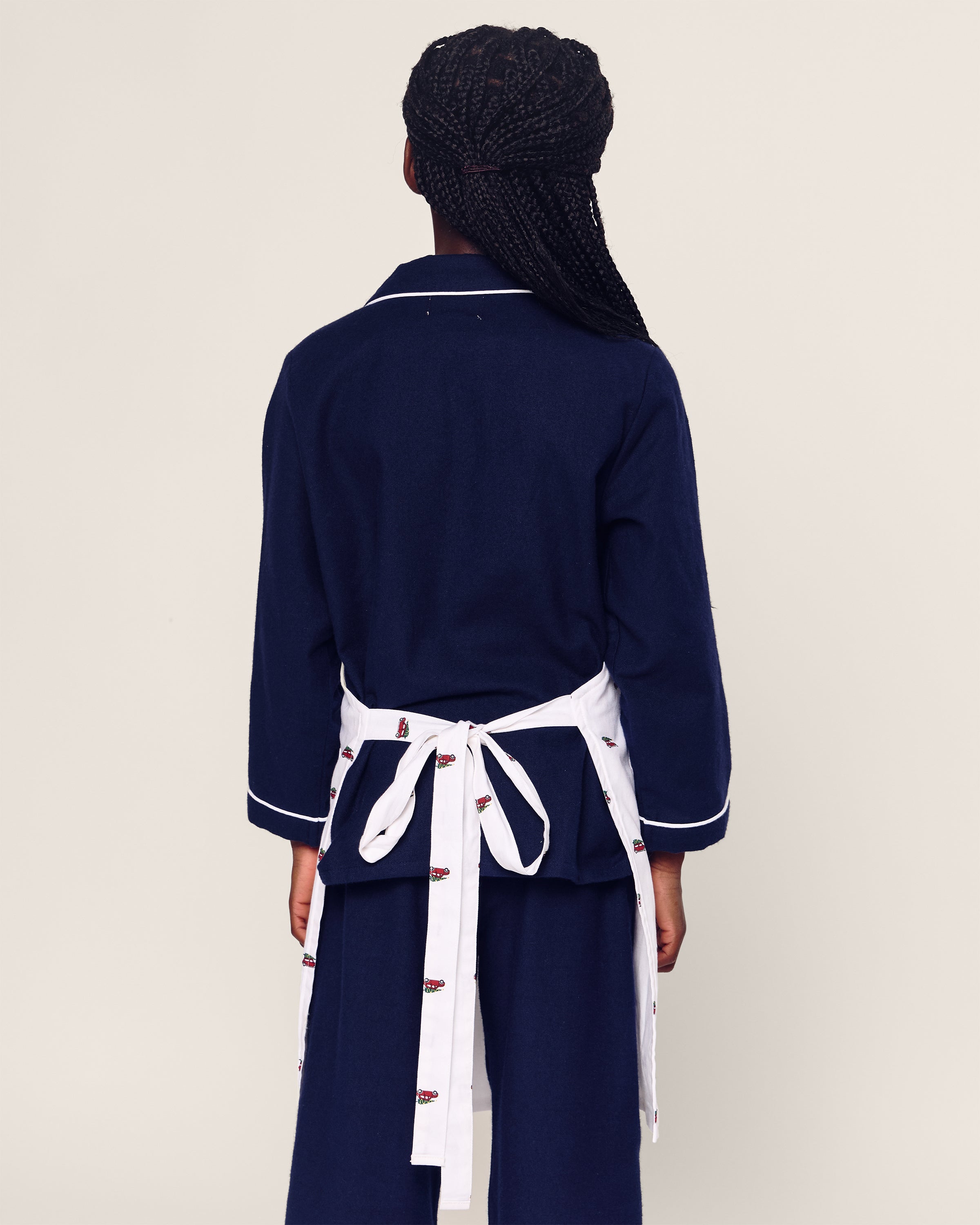 A person with long braided hair, wearing Petite Plumes dark blue long-sleeve outfit with white trim and the Kids Apron in Holiday Journey tied at the back, stands facing away against a plain light background—a perfect ensemble for holiday baking.