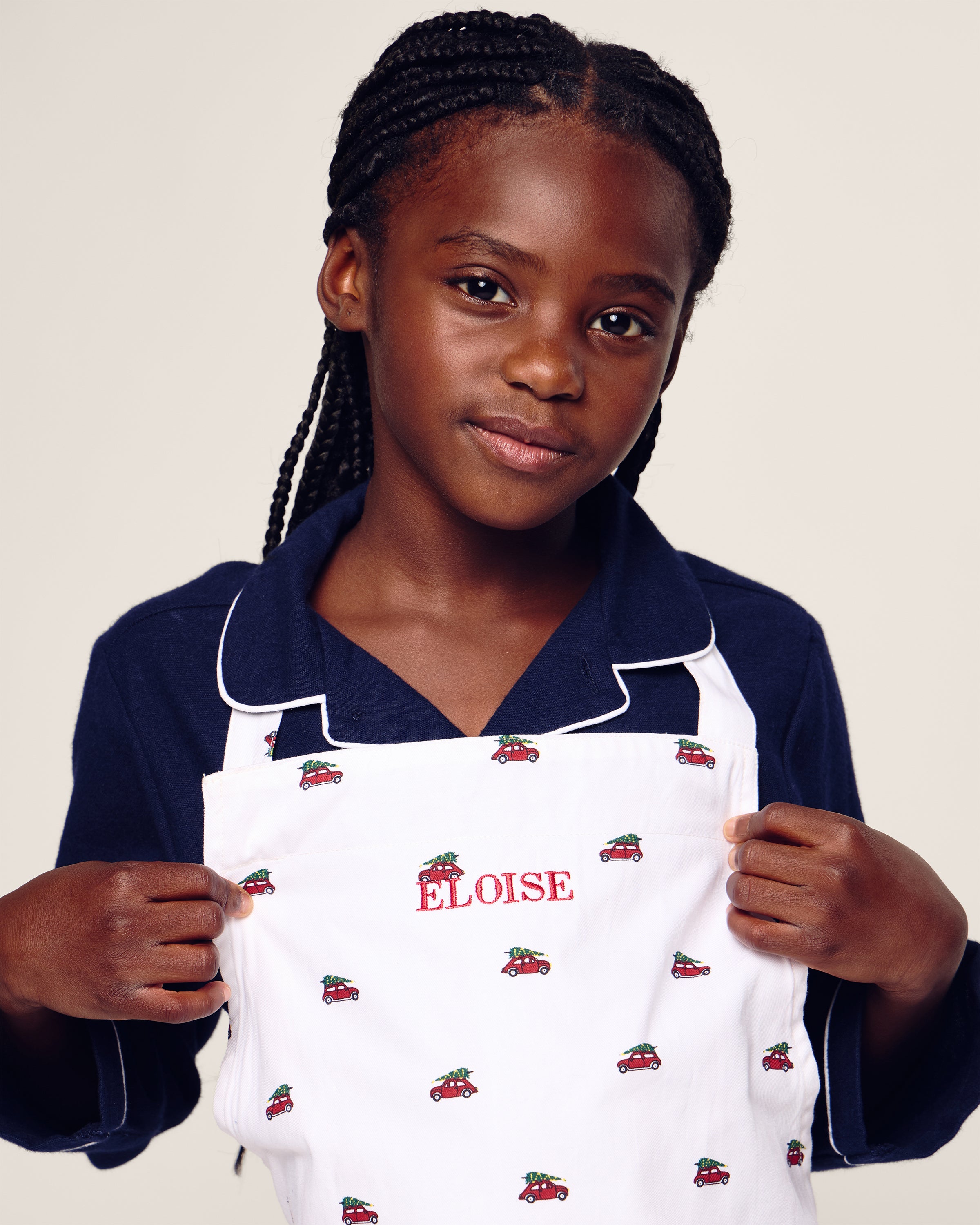 A young girl with braided hair wears Petite Plumes Kids Apron in Holiday Journey, a navy shirt, and a white apron with car designs. ELOISE is written in red on the apron. She faces the camera with a neutral expression, making it ideal for holiday baking in childrens sizes.