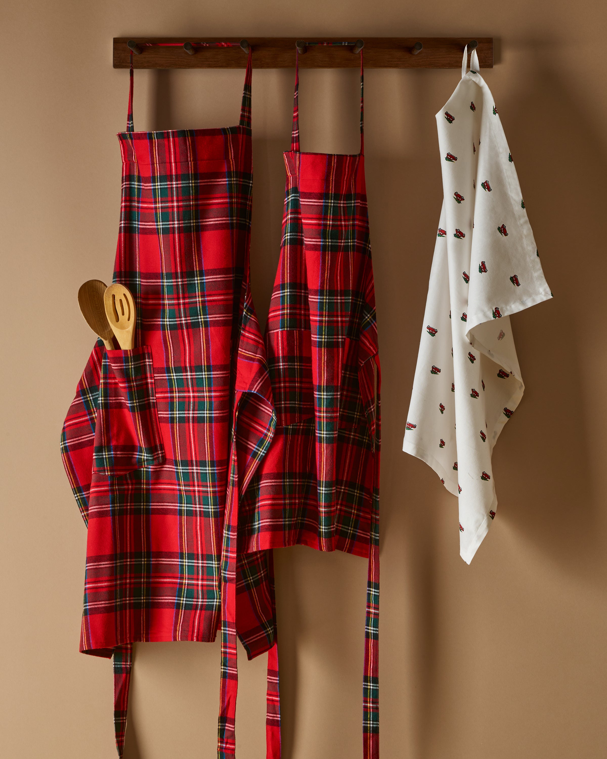 Two Petite Plume Kids Aprons in Imperial Tartan with pockets hang on a wooden peg rack, alongside a white dish towel with red and black patterns. A wooden spoon and spatula peek from one apron pocket, perfect for holiday baking against a light brown backdrop.