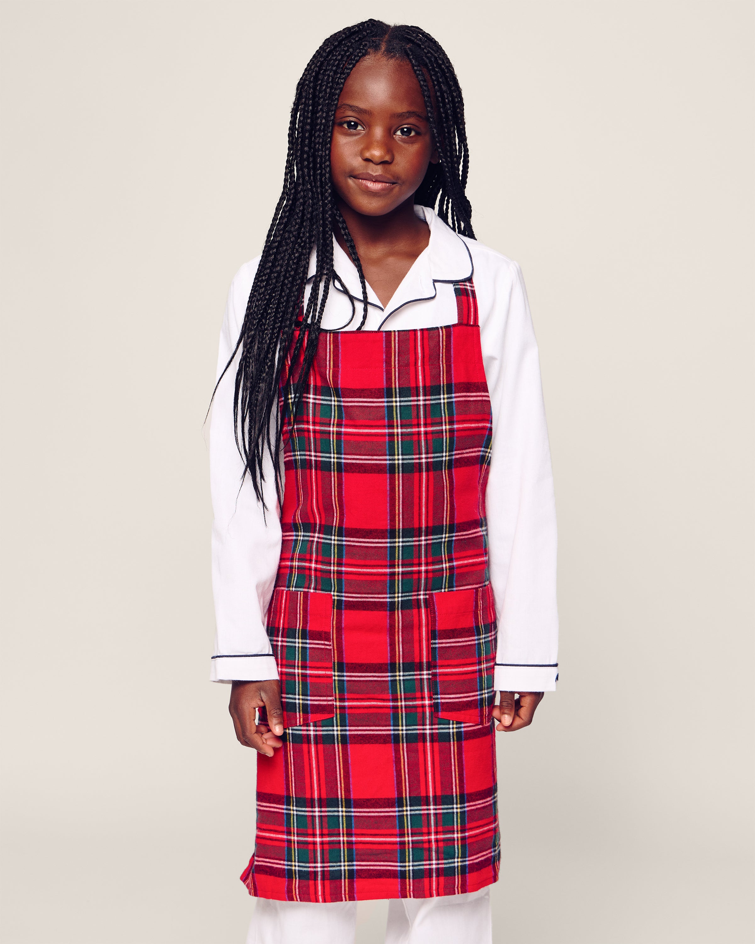 A young girl with long braided hair dons Petite Plumes Kids Apron in Imperial Tartan over her white pajamas. She stands neutral against a plain backdrop, ready for culinary adventures.