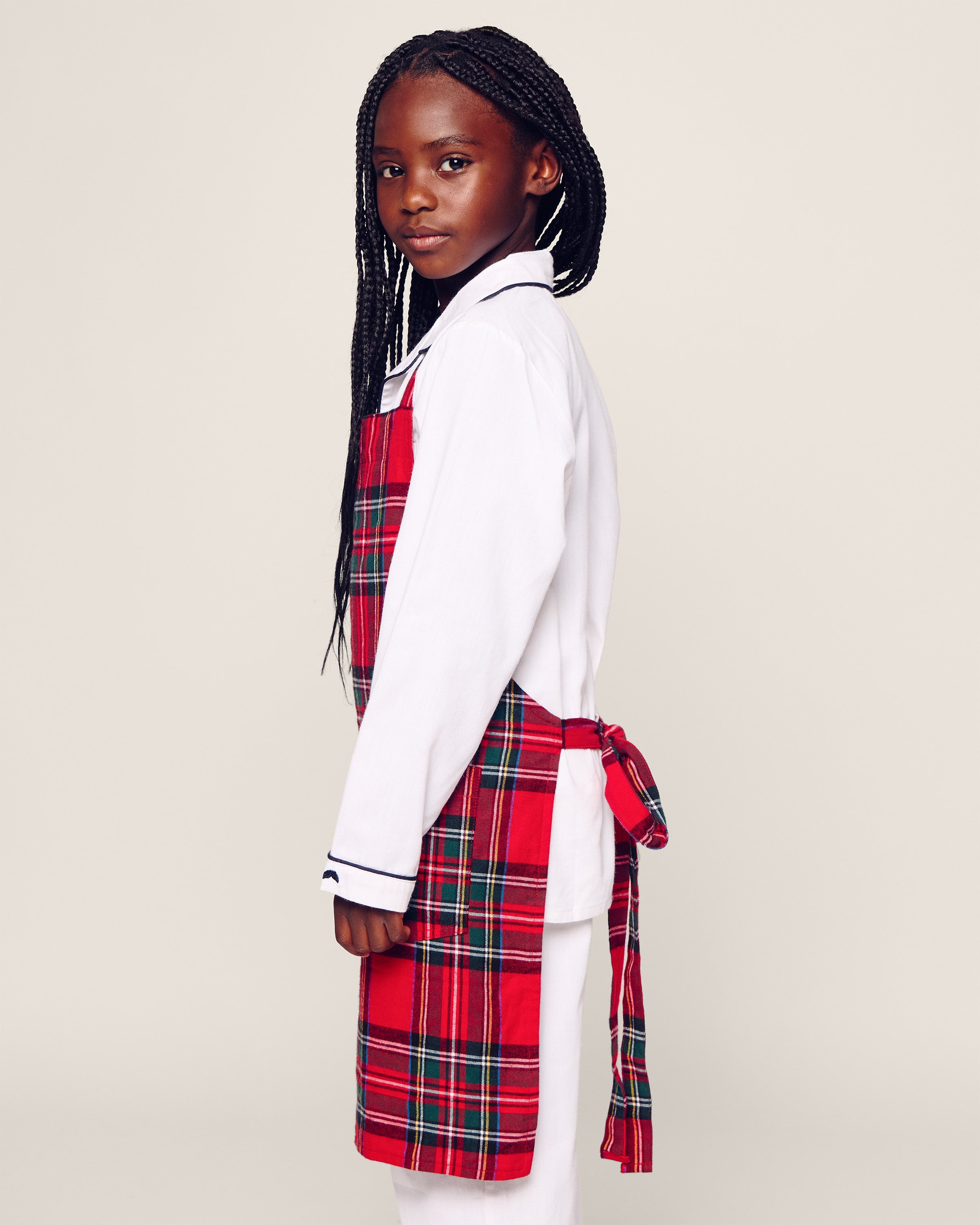 A young girl with long braided hair stands sideways, wearing a white shirt and the Petite Plume Kids Apron in Imperial Tartan, tied at the back. She gazes confidently at the camera against a plain background.