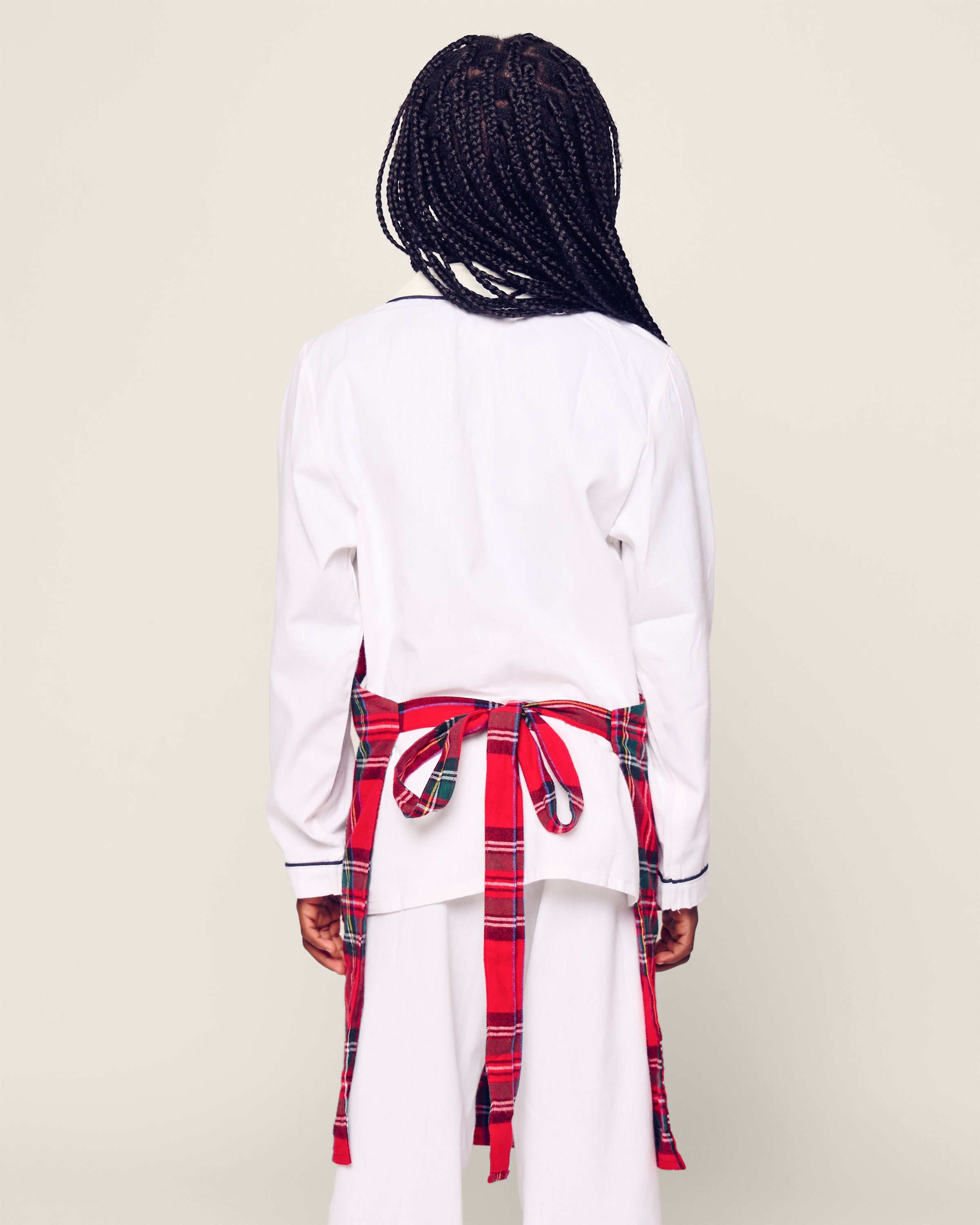 Wearing braided hair and a white long-sleeve top, this person stands against a neutral backdrop, flaunting Petite Plumes Kids Apron in Imperial Tartan tied around their waist like a festive holiday apron.