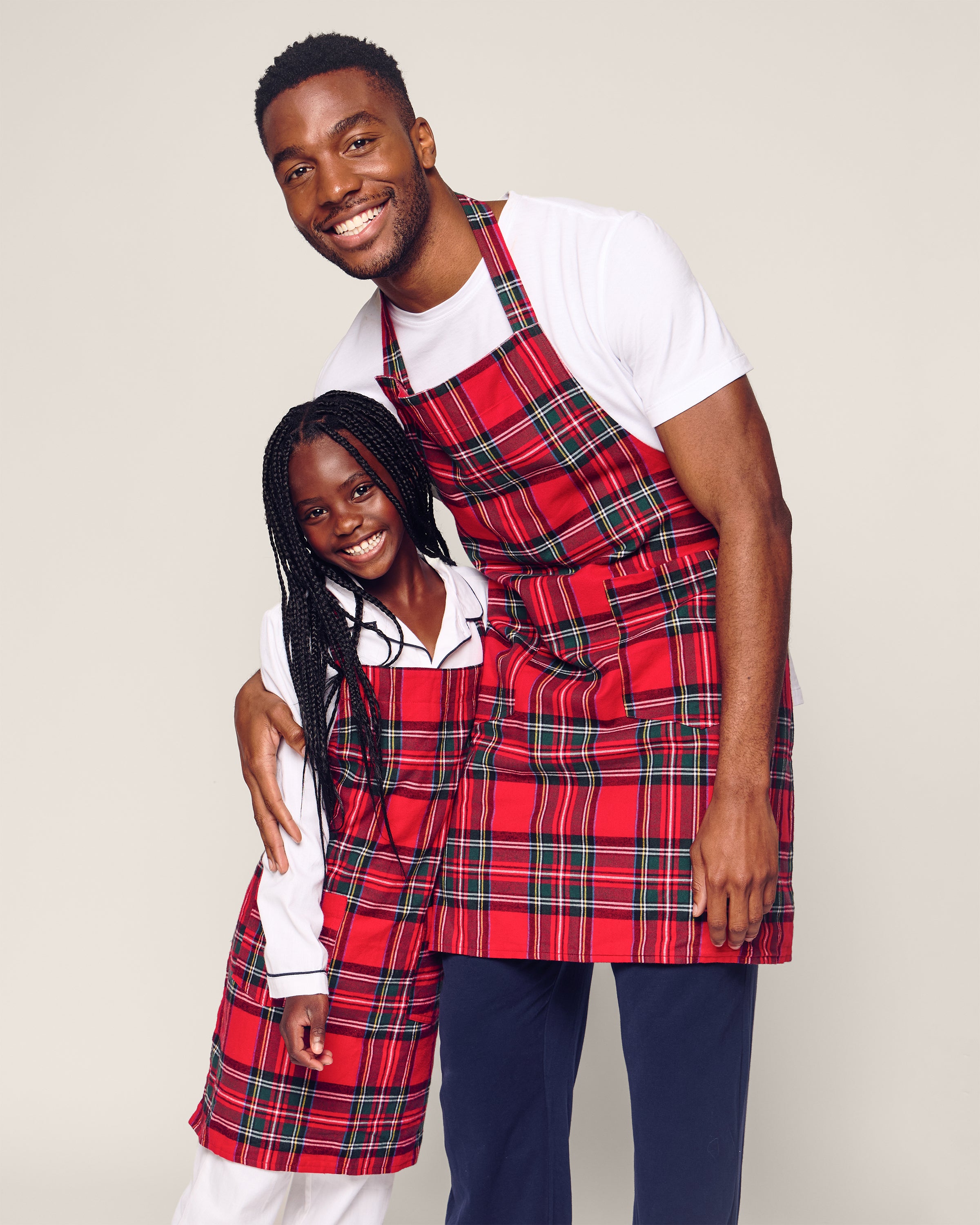 A man in a white tee and dark pants, and a girl in a white long-sleeve top are smiling joyfully. The duo showcases their holiday cheer in festive red plaid baking aprons, including the girls Kids Apron in Imperial Tartan by Petite Plume, against a plain light-colored background.