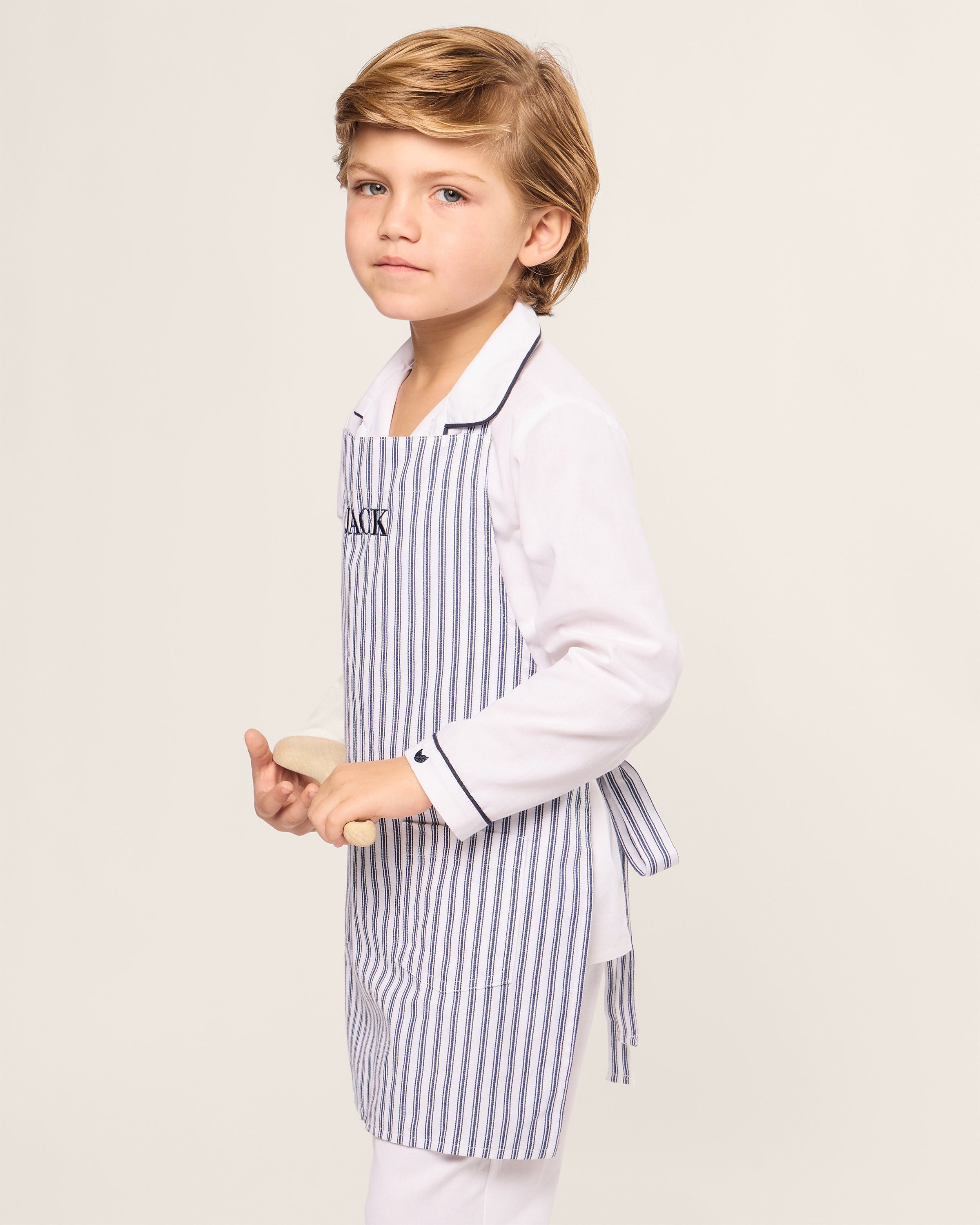 A young child with light brown hair, wearing a Petite Plume Kids Apron in Navy French Ticking and holding a small rolling pin, stands gazing ahead with a neutral expression, ready for holiday baking against a plain backdrop.