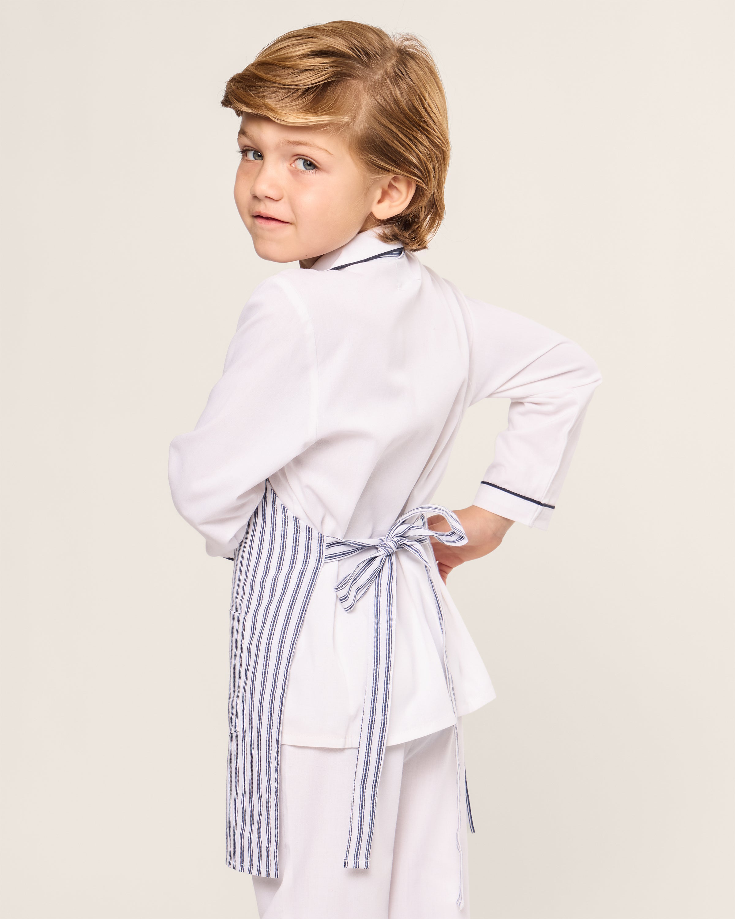 A young child with light brown hair wears a white top and the Navy French Ticking Kids Apron by Petite Plume, ideal for holiday baking. They stand with their back to the camera, turning their head to the left, against a plain background.