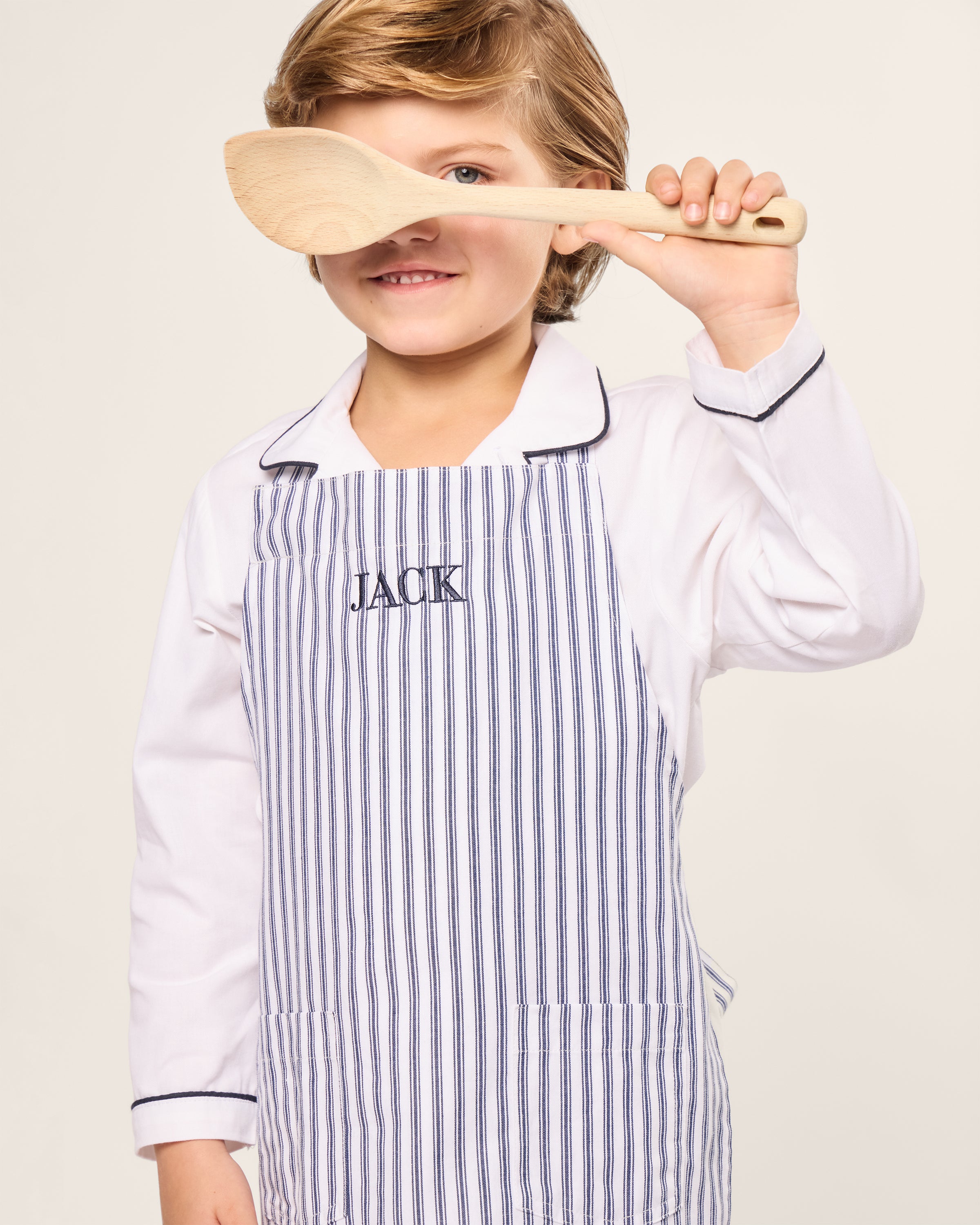 A child joyfully engages in holiday baking, wearing a Petite Plume Kids Apron in Navy French Ticking with Jack embroidered on it. Holding a wooden spoon playfully, the child smiles warmly in a white shirt with dark piping against a plain, light background.