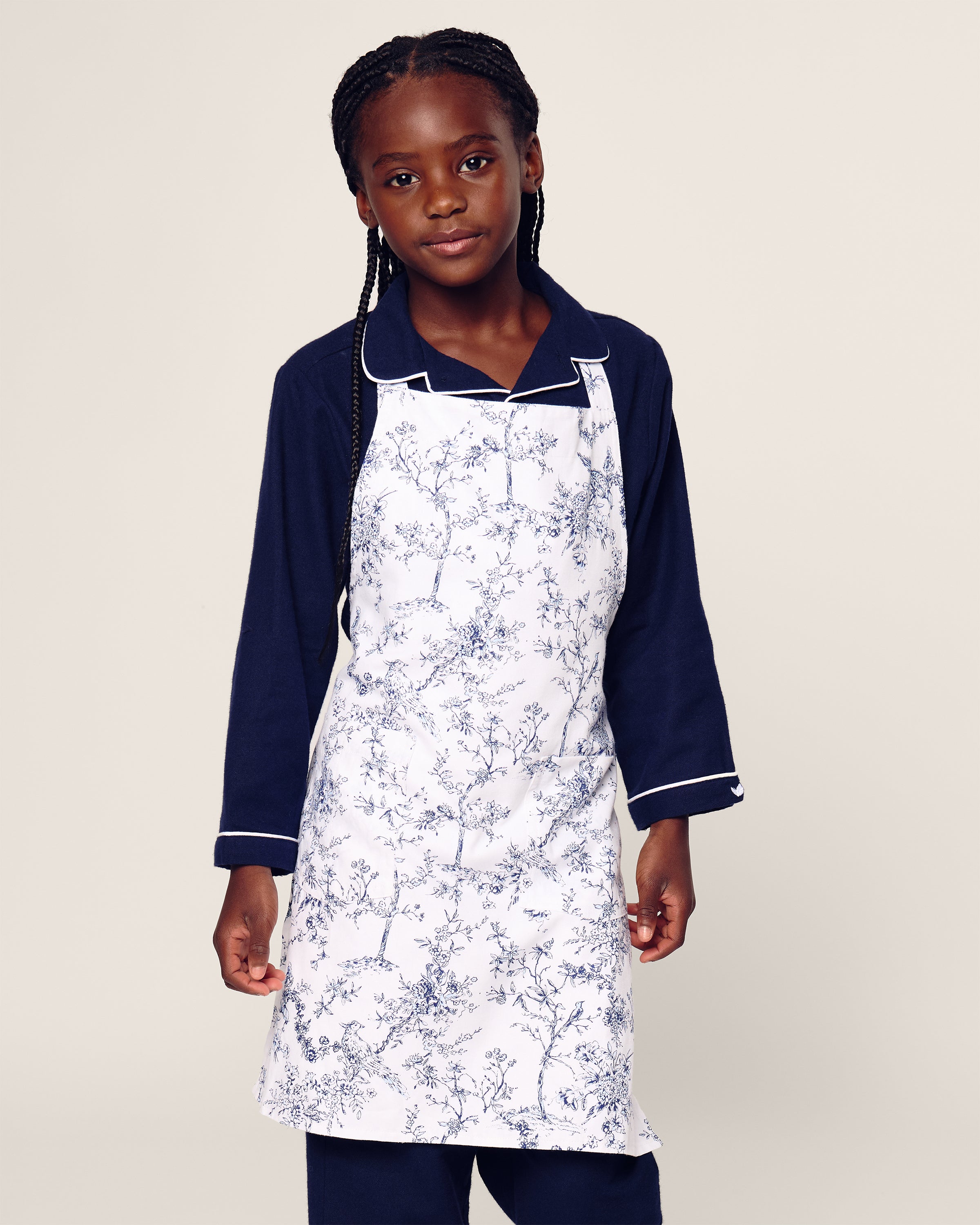 A child in a Petite Plume blue long-sleeve shirt and a Kids Apron in Timeless Toile stands facing forward against a plain backdrop, prepared for delightful holiday baking.