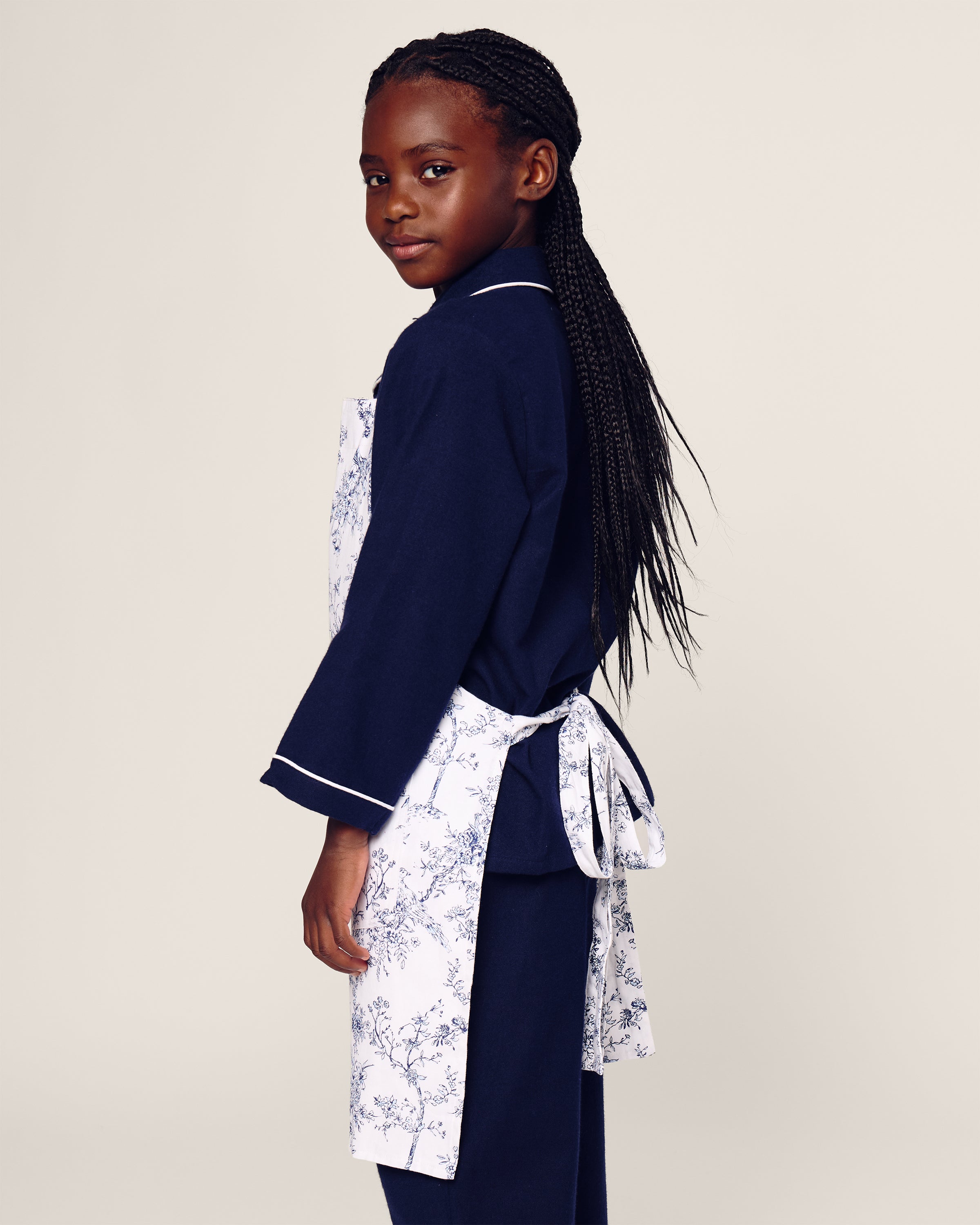 A young girl with long braids stands sideways, exuding botanical elegance in a navy blue outfit and Petite Plumes Kids Apron in Timeless Toile, featuring a floral pattern tied at the back. She confidently faces the camera against a plain backdrop, ready for her holiday baking adventure.