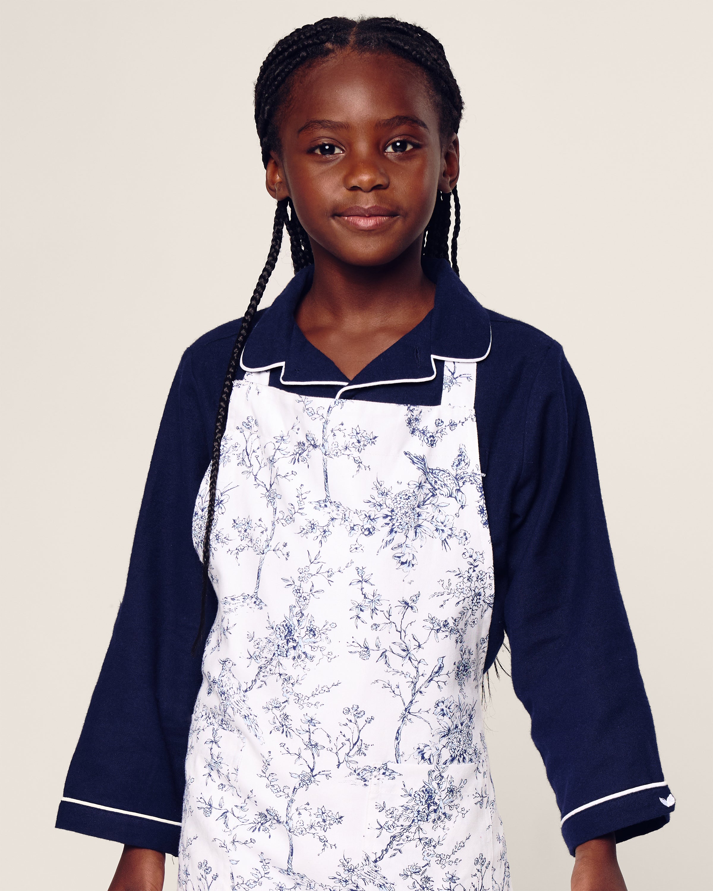 A young girl embodies botanical elegance in a navy blue shirt with white piping and Petite Plumes Timeless Toile Kids Apron. With braided hair and a neutral expression, she stands against a plain background, capturing the charm of holiday baking.