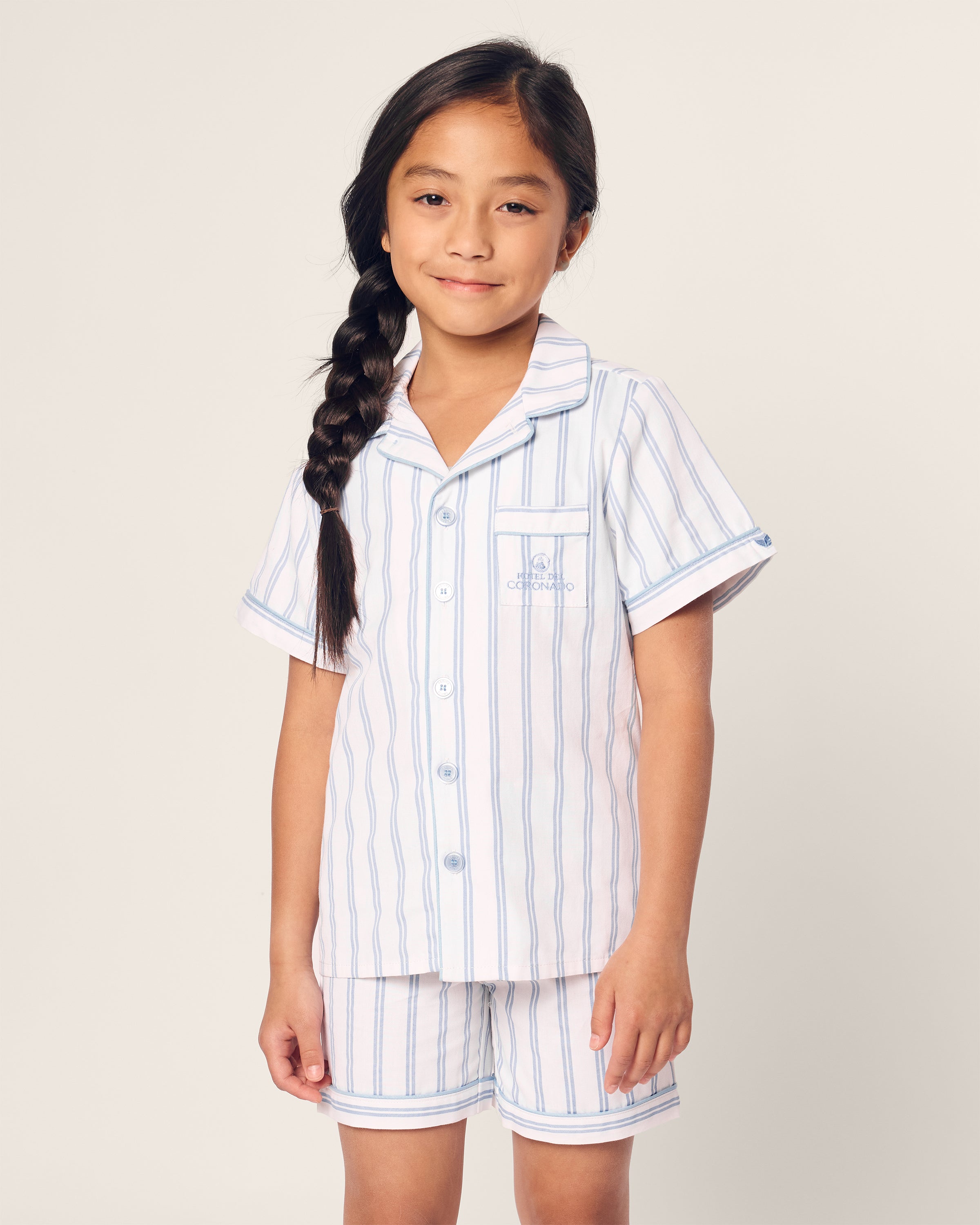 A young girl wearing the Kids Twill Periwinkle Stripe Short Set Hotel del Coronado x Petite Plume from Petite Plume stands against a plain background. Her long, dark hair is in a side braid as she gently smiles at the camera, capturing the timeless charm reminiscent of Hotel del Coronado.