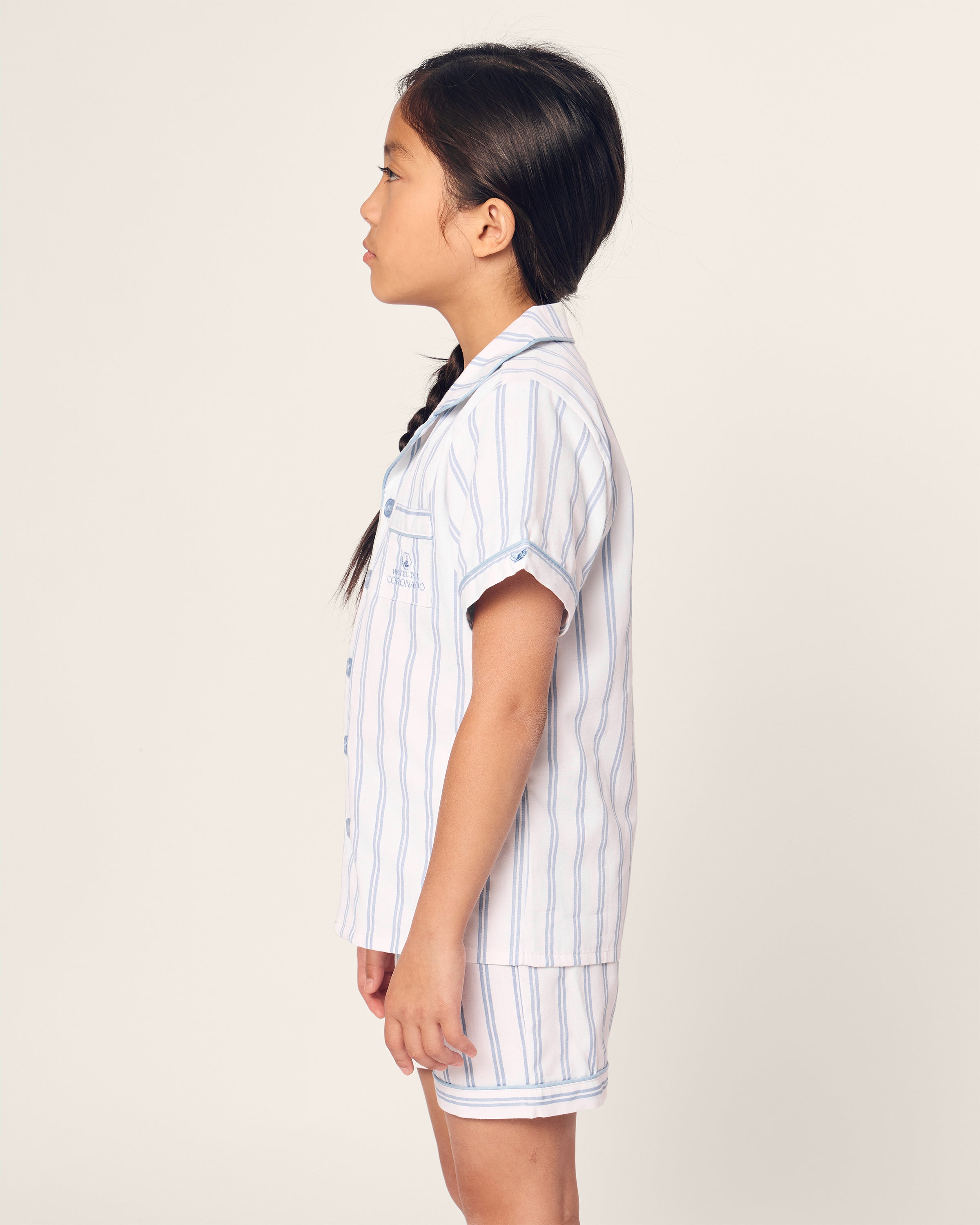 A young girl with long dark hair in a braid stands sideways, wearing the limited-edition Kids Twill Periwinkle Stripe Short Set by Hotel del Coronado x Petite Plume. This classic pajama set features short sleeves and light blue and white vertical stripes with buttons down the front.