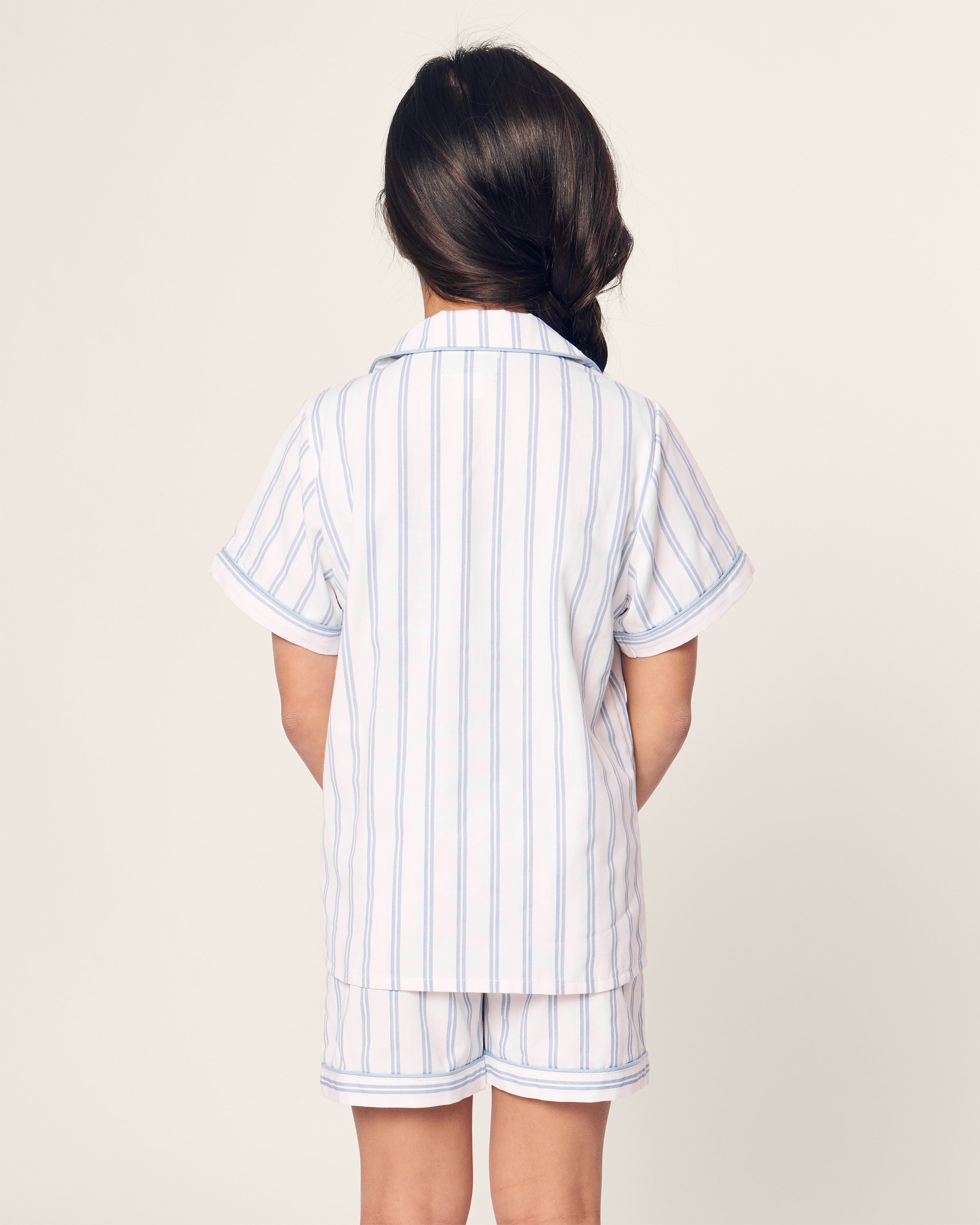 A child with long dark hair in a braid stands facing away, wearing a Petite Plume Kids Twill Periwinkle Stripe Short Set from the Hotel del Coronado collaboration. The pajama set includes a short-sleeved button-up shirt and shorts with light blue and white stripes, against a simple, light-colored backdrop.