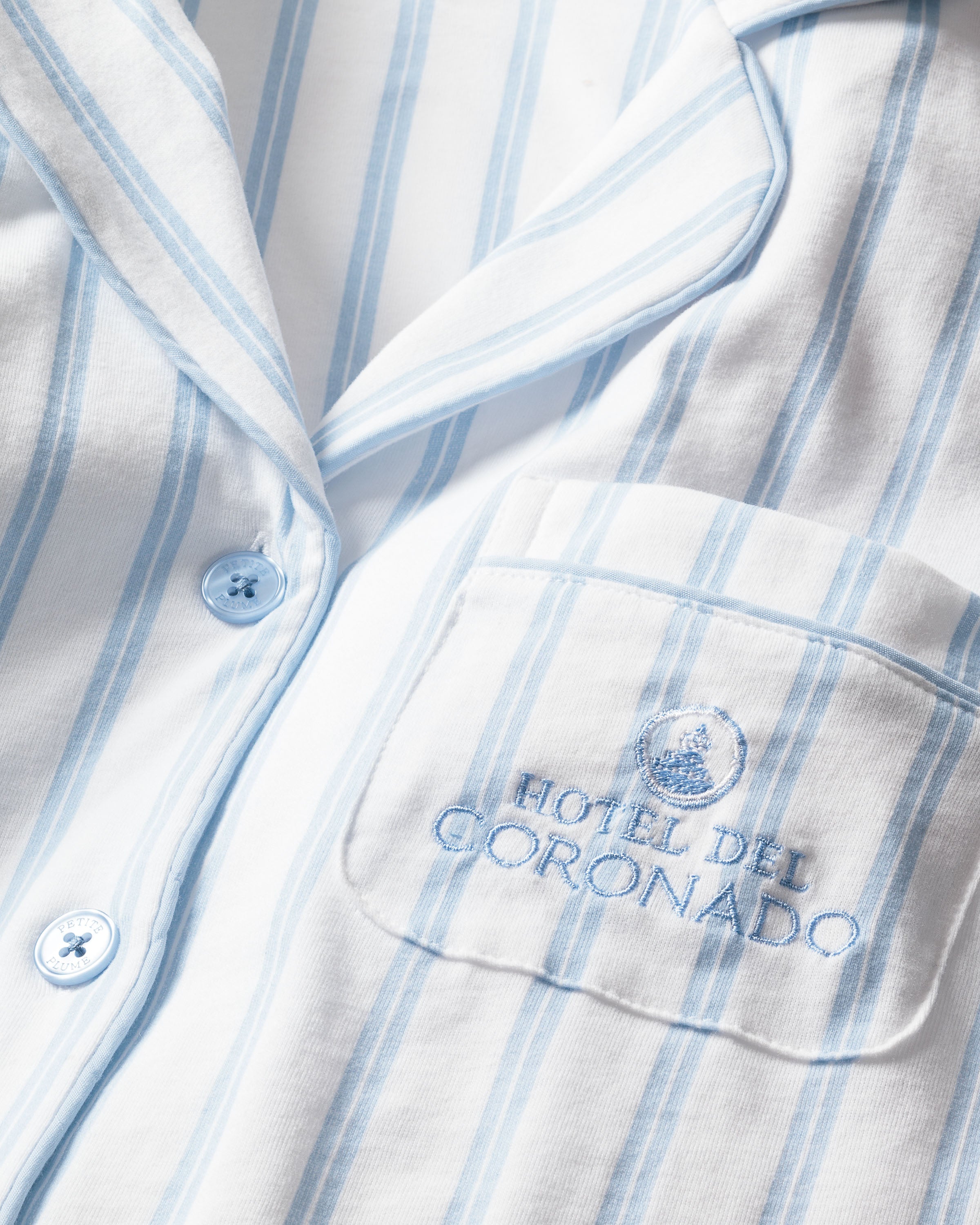 Close-up of the Kids Twill Periwinkle Stripe Short Set by Petite Plume x Hotel del Coronado. This limited-edition pajama top features light blue vertical stripes, a chest pocket embroidered with HOTEL DEL CORONADO and emblem, light blue buttons, and a visible collar.