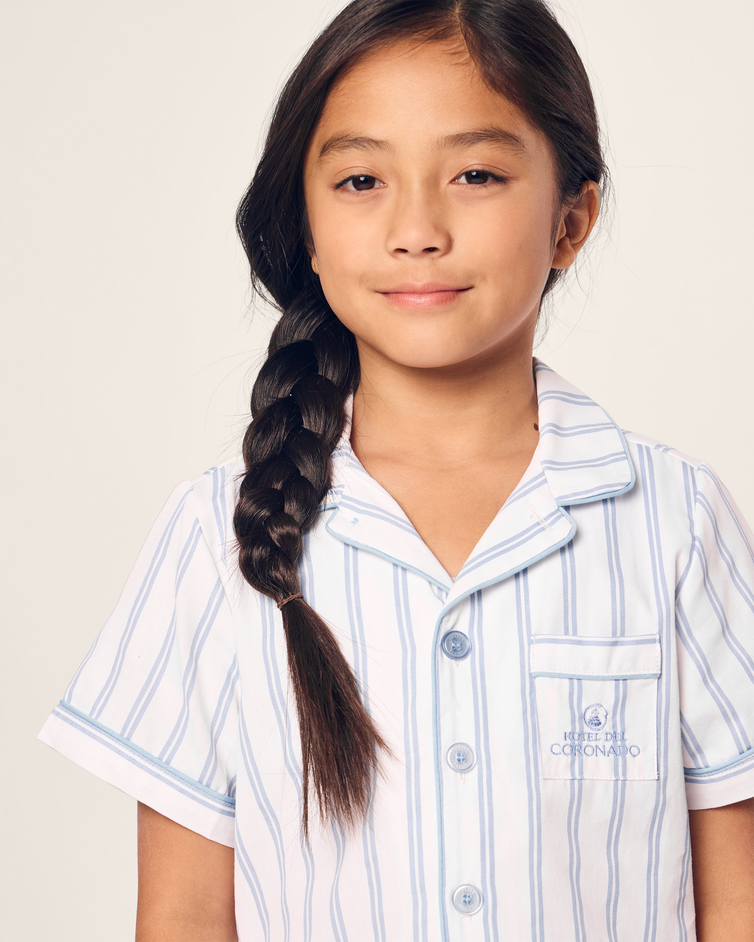 A young girl with long dark hair in a braid smiles softly, wearing the Kids Twill Periwinkle Stripe Short Set Hotel del Coronado x Petite Plume. The light blue and white striped design, with short sleeves and a collar, perfectly complements the neutral background.