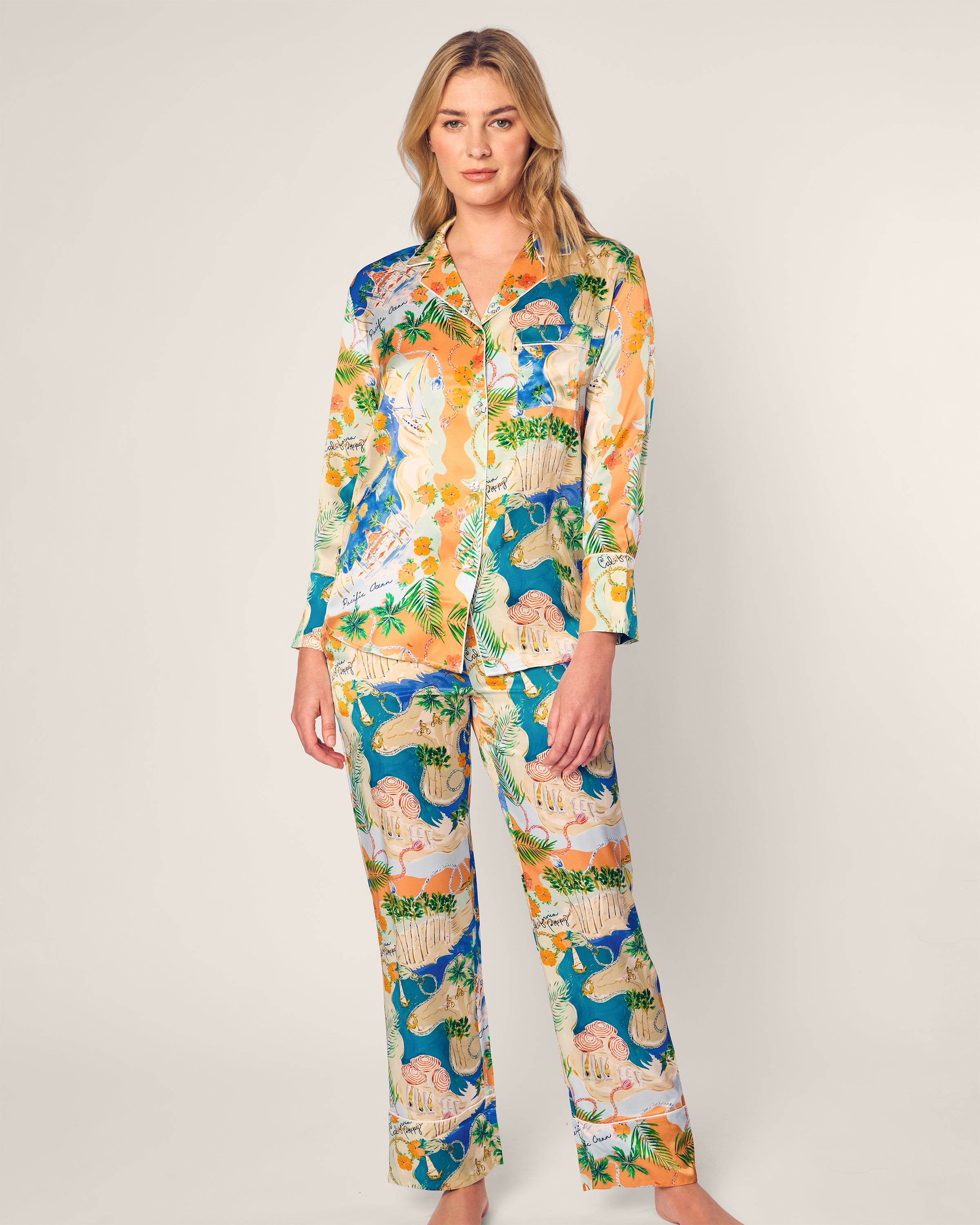 A person models the Womens Silk Print Pajama Set Hotel del Coronado x Petite Plume by Petite Plume against a plain background. The luxury sleepwear, made of colorful pure silk featuring animals and plants, includes a button-up top and loose, full-length pants.