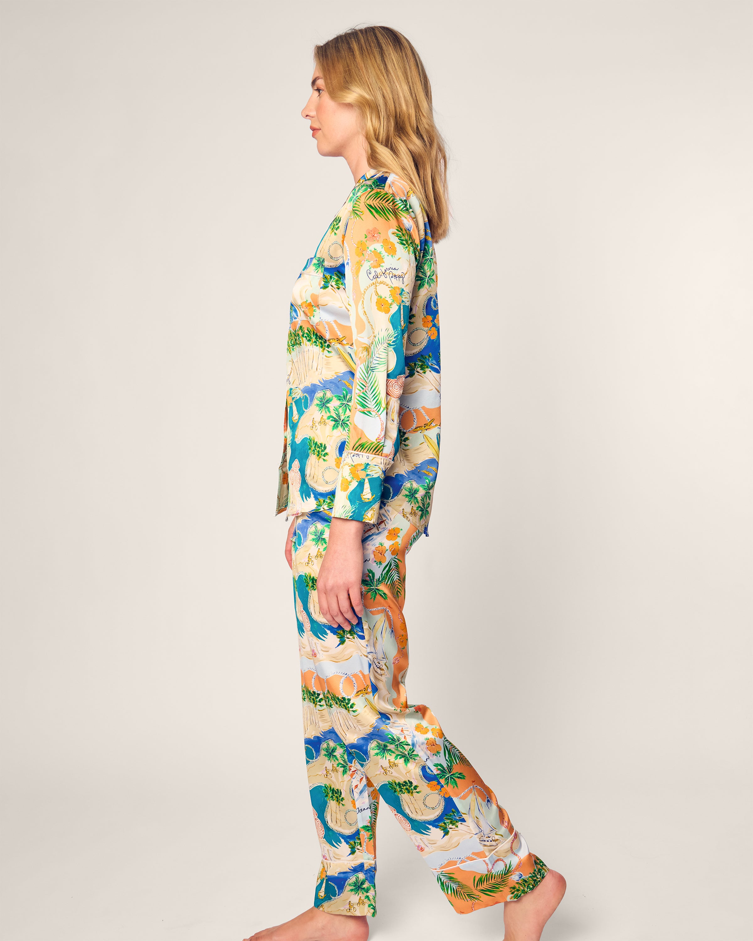 A person with long blonde hair stands barefoot in side profile, wearing the Womens Silk Print Pajama Set by Petite Plume x Hotel del Coronado, featuring green, blue, orange, and white leaf and flower prints on a light gray background.