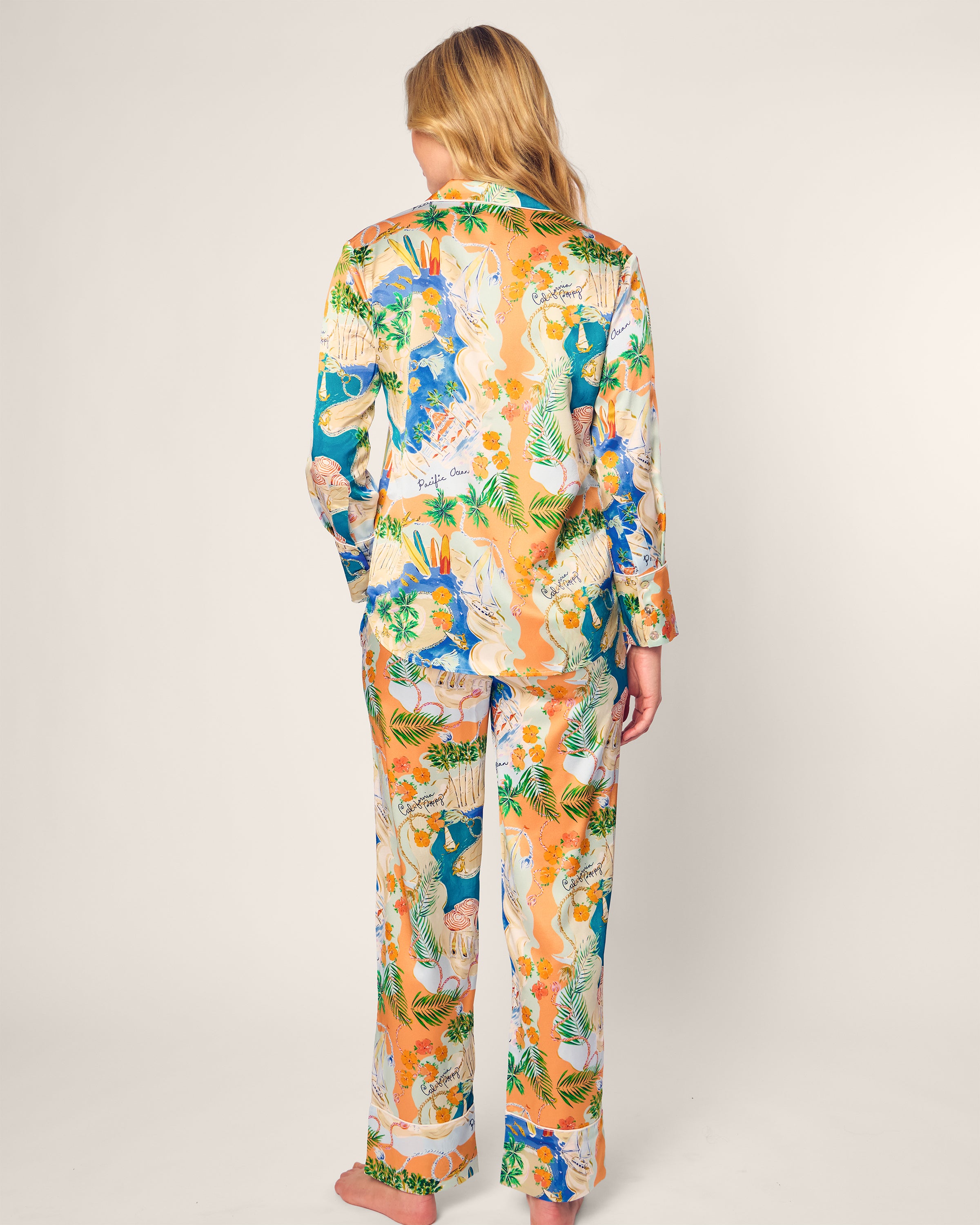 A person in the Womens Silk Print Pajama Set by Petite Plume x Hotel del Coronado, featuring a long-sleeved top and pants with an intricate animal and foliage pattern in vibrant orange, green, and blue shades. The person stands barefoot against a neutral background.