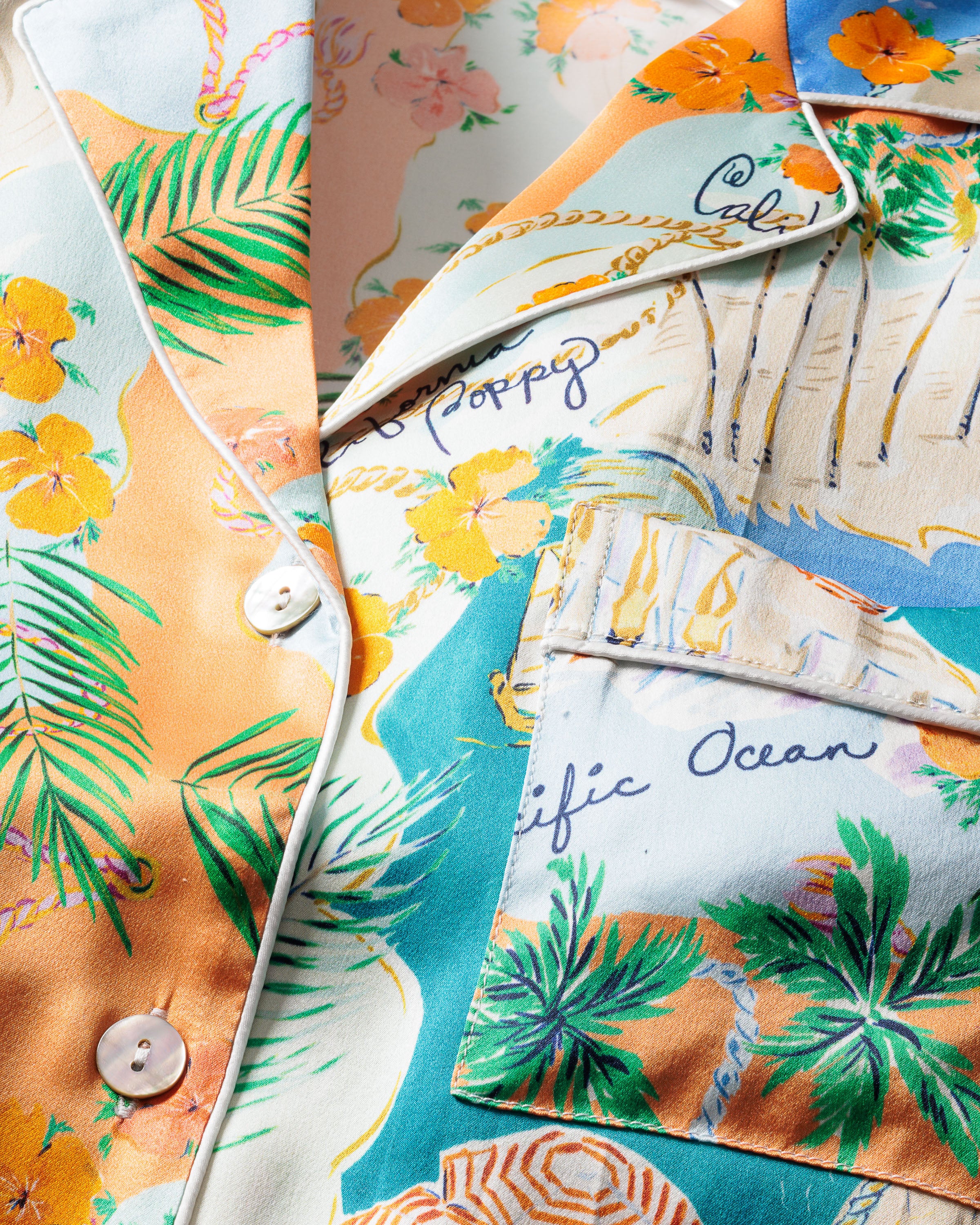 Close-up of the Womens Silk Print Pajama Set from the Hotel del Coronado x Petite Plume collection. This luxury sleepwear features a colorful tropical theme with palm fronds, flowers, oceanic imagery, white buttons, and text like Pacific Ocean and Poppy.