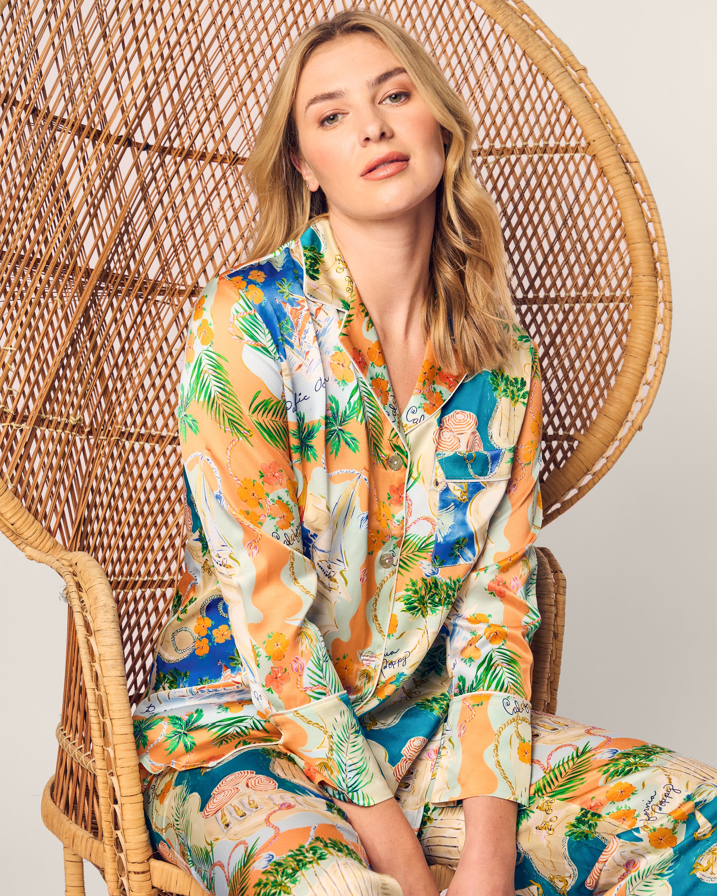 Sitting on a wicker chair, they exude elegance in a Womens Silk Print Pajama Set from the Hotel del Coronado x Petite Plume collection. With long, wavy blond hair and a relaxed expression, the light-colored background completes the scene.