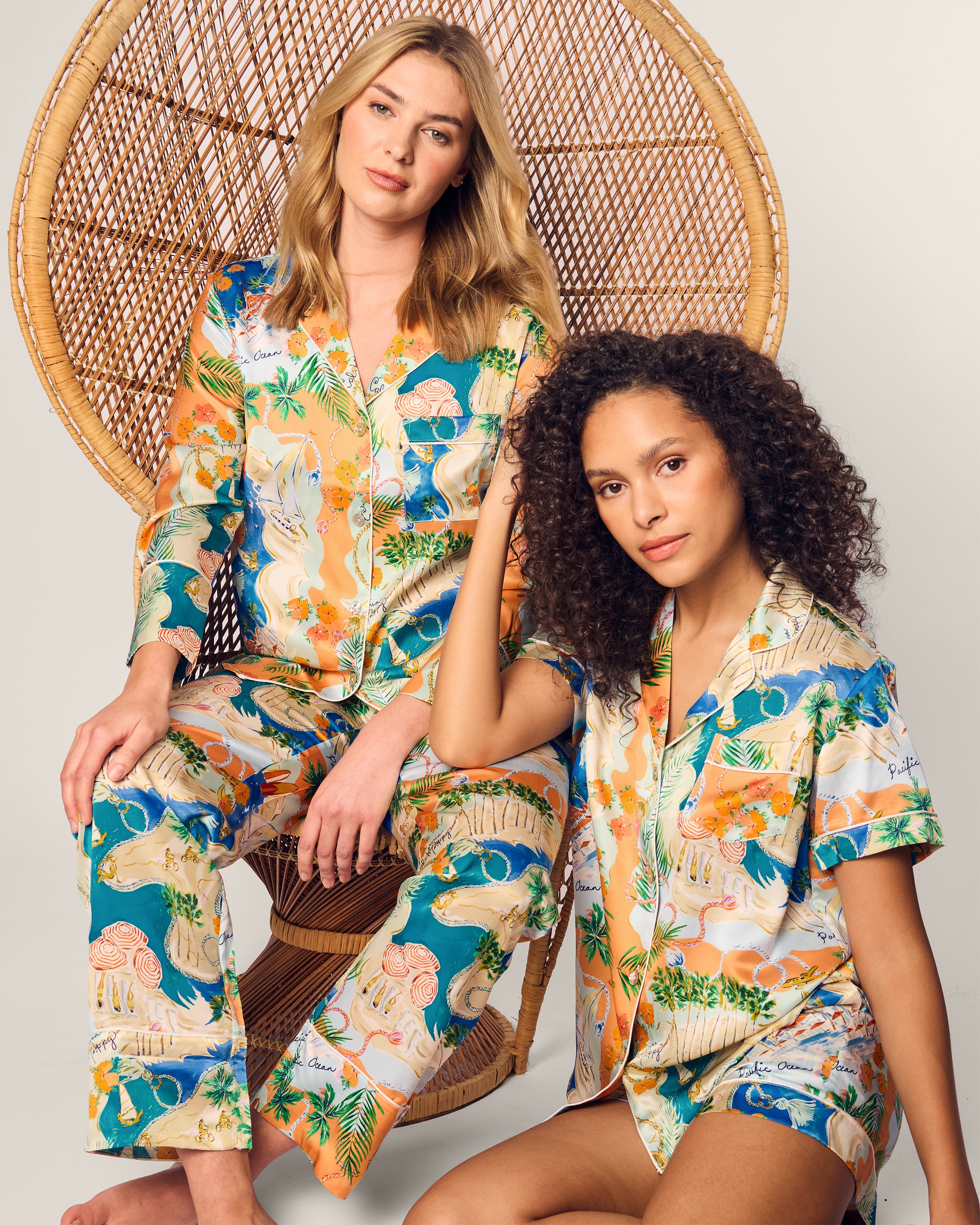 Two women relax on a wicker chair in matching Womens Silk Print Pajama Sets by Hotel del Coronado x Petite Plume. The sets vibrant animal and nature patterns bring elegance to the serene, neutral backdrop—one woman sits while the other leans against the chair.