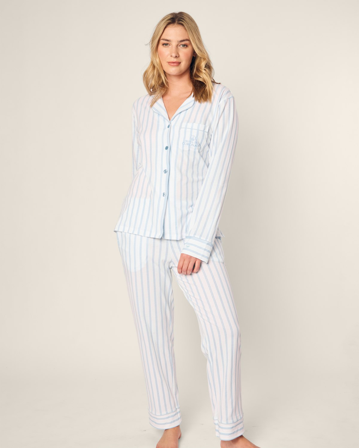 A woman dons the Womens Pima Pajama Set by Petite Plume, part of the Hotel del Coronado x Petite Plume collection. Made from soft Peruvian Pima cotton, the light blue and white striped set includes a long-sleeved buttoned top with a chest pocket and full-length pants. She looks slightly to the side against a plain background.