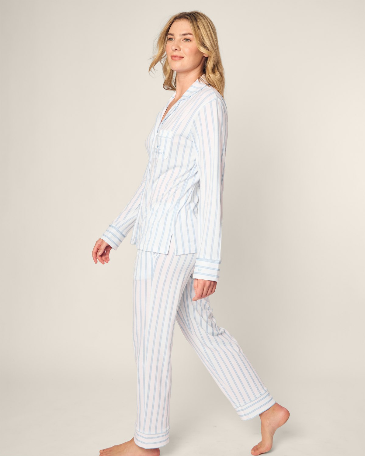 A person barefoot on a plain background wears the Hotel del Coronado x Petite Plume Womens Pima Pajama Set by Petite Plume, crafted from luxurious yarn-dyed Peruvian Pima cotton in light blue and white stripes, featuring a classic button-up top and long pants. They appear relaxed.