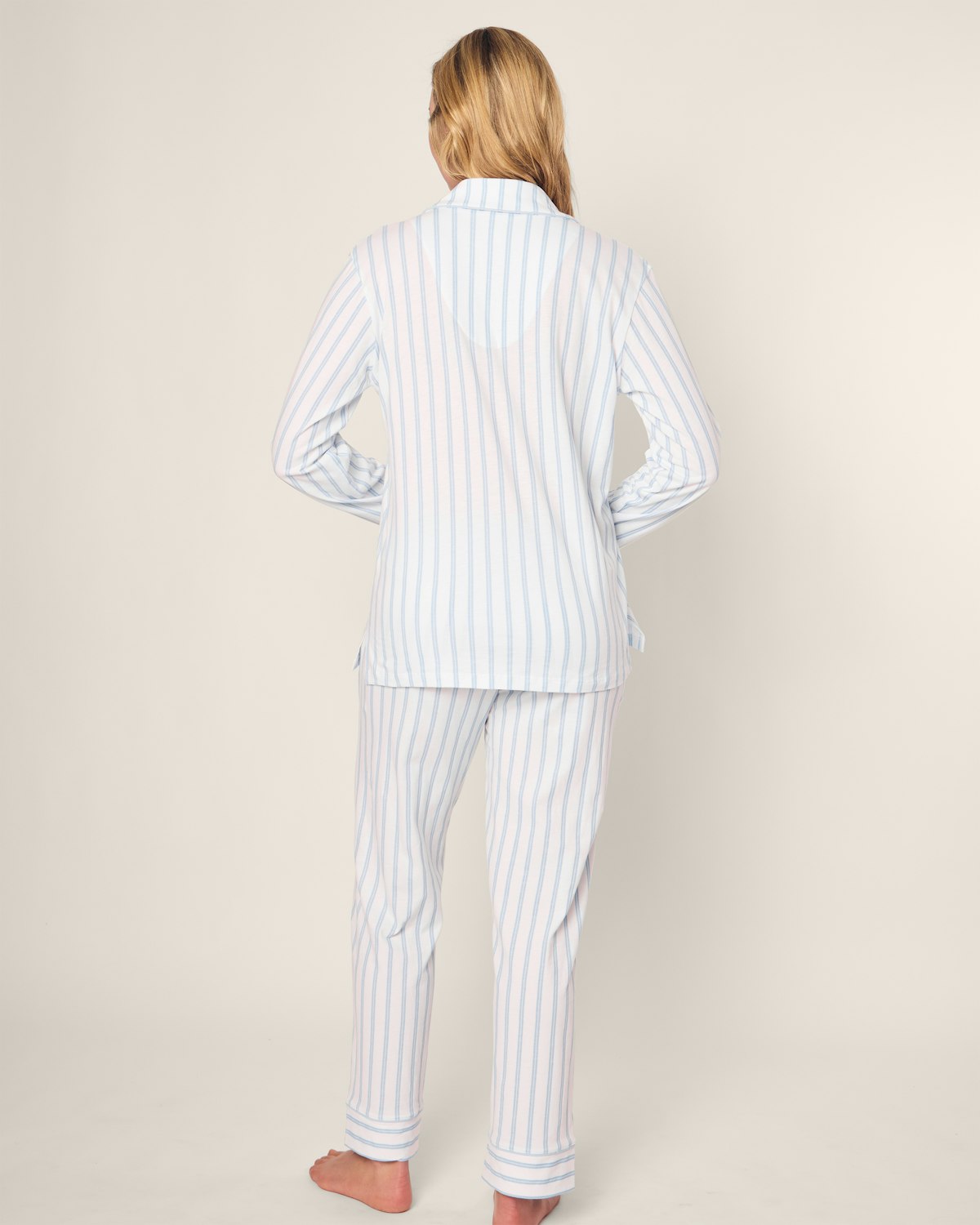 A person with long blonde hair stands barefoot, facing away in luxurious Womens Pima Pajama Set Hotel del Coronado x Petite Plume by Petite Plume. This light blue and white vertically striped set includes a yarn-dyed long-sleeved top and pants, against a plain light background.