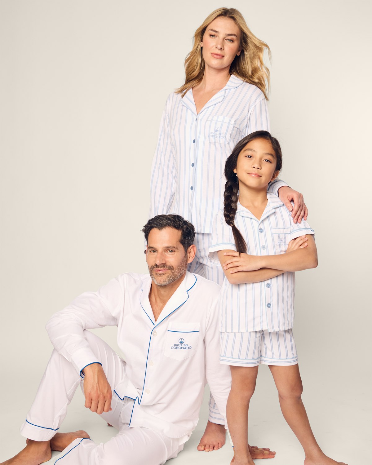 A family of three poses against a plain backdrop in matching yarn-dyed striped pajamas made from Peruvian Pima cotton. The man kneels, the woman (wearing Womens Pima Pajama Set Hotel del Coronado x Petite Plume by Petite Plume) stands behind him, and a young girl stands next to them, all appearing relaxed and content.