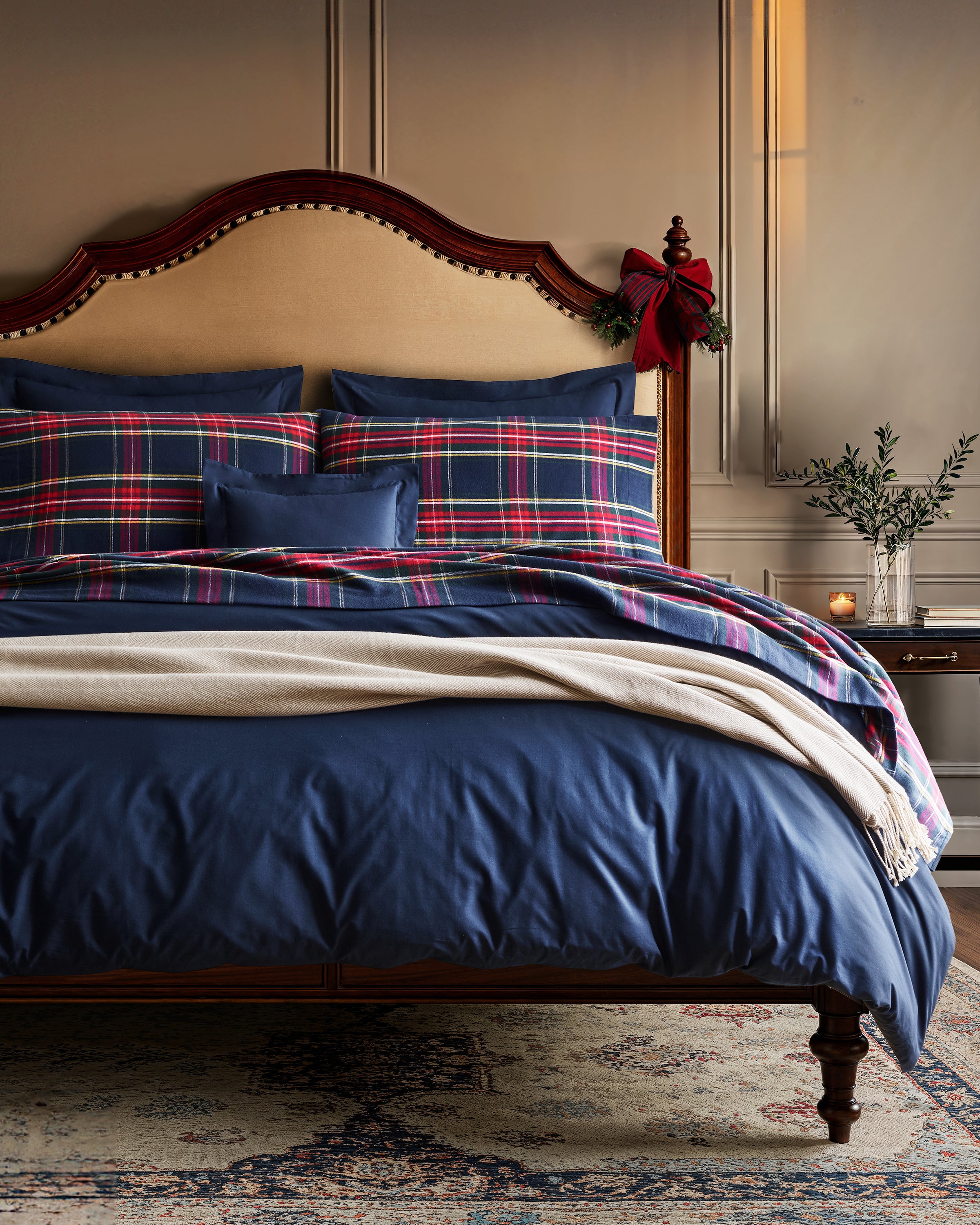 Elegant bedroom featuring a wooden bed with blue plaid bedding and the Herringbone Throw in Beige by Petite Plume, accented by a festive red bow. A small table with a vase of greenery and a lit candle enhances the cozy home decor, creating a warm, inviting ambiance.