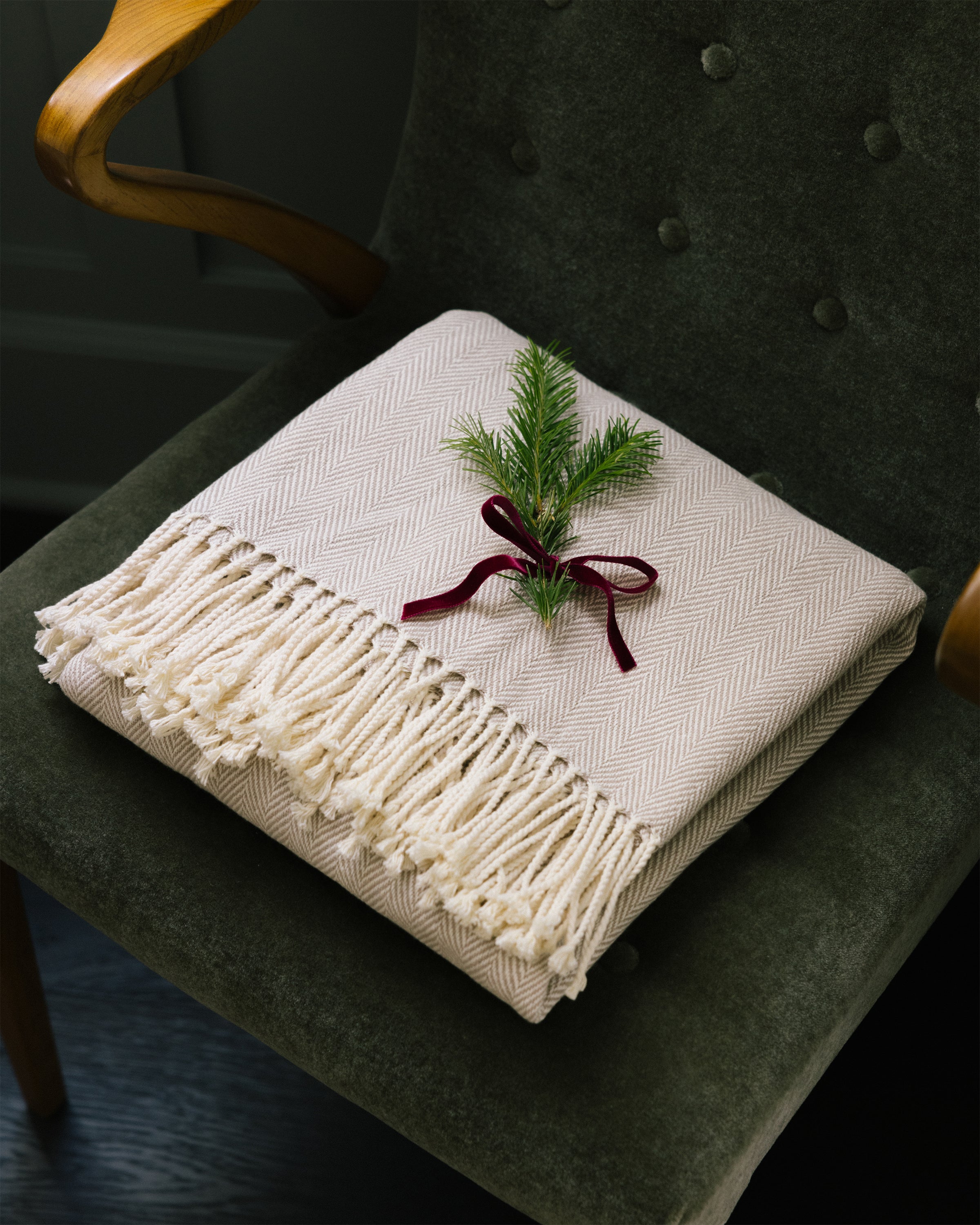 A folded Herringbone Throw in Beige by Petite Plume with fringe rests on a green upholstered chair. Its tied with a burgundy ribbon and adorned with green pine sprigs, creating charming home decor.