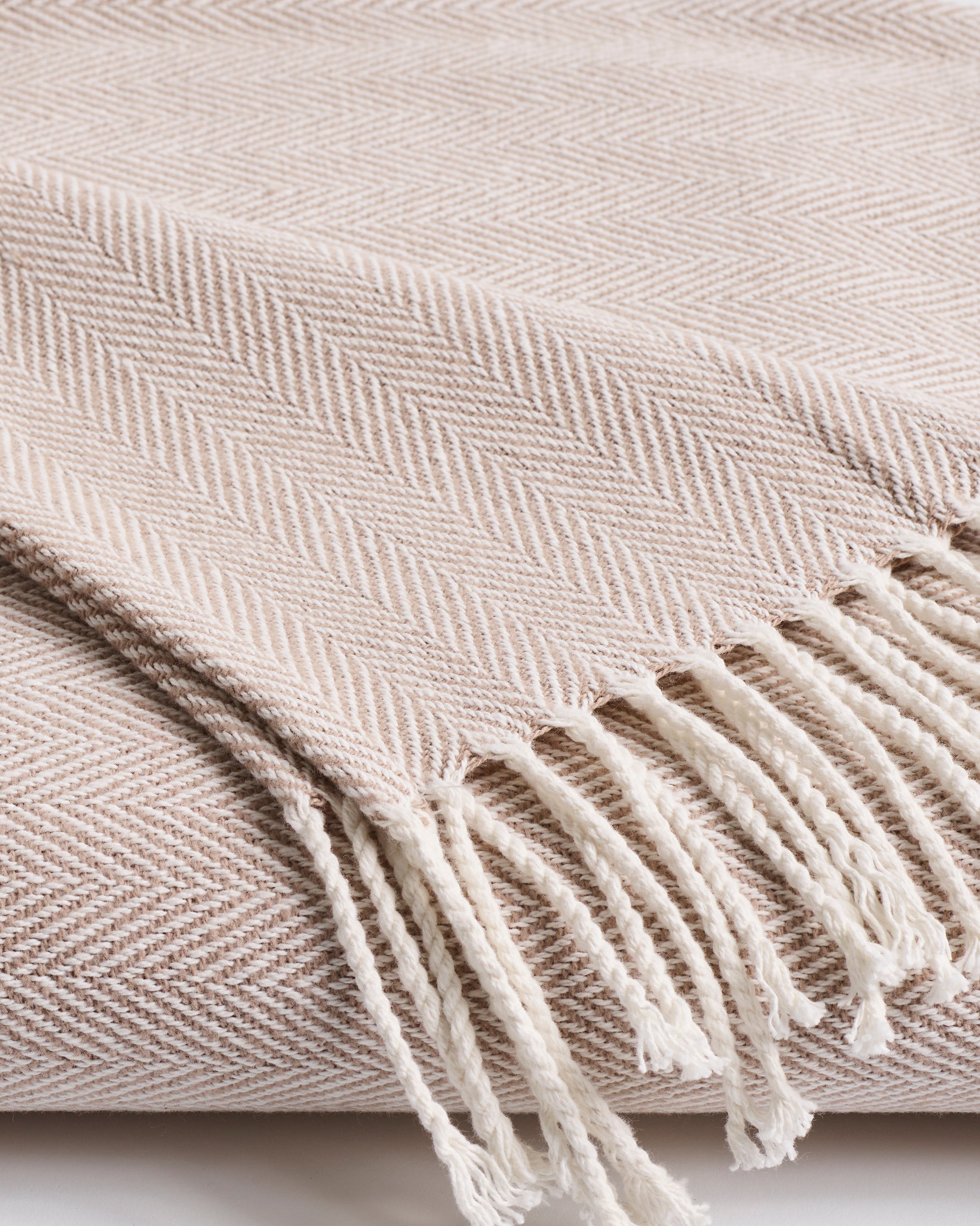 Close-up of the Petite Plume Herringbone Throw in Beige, made from soft cotton. It features white fringe tassels on one edge, adding texture and coziness, making it a perfect addition to any home decor.