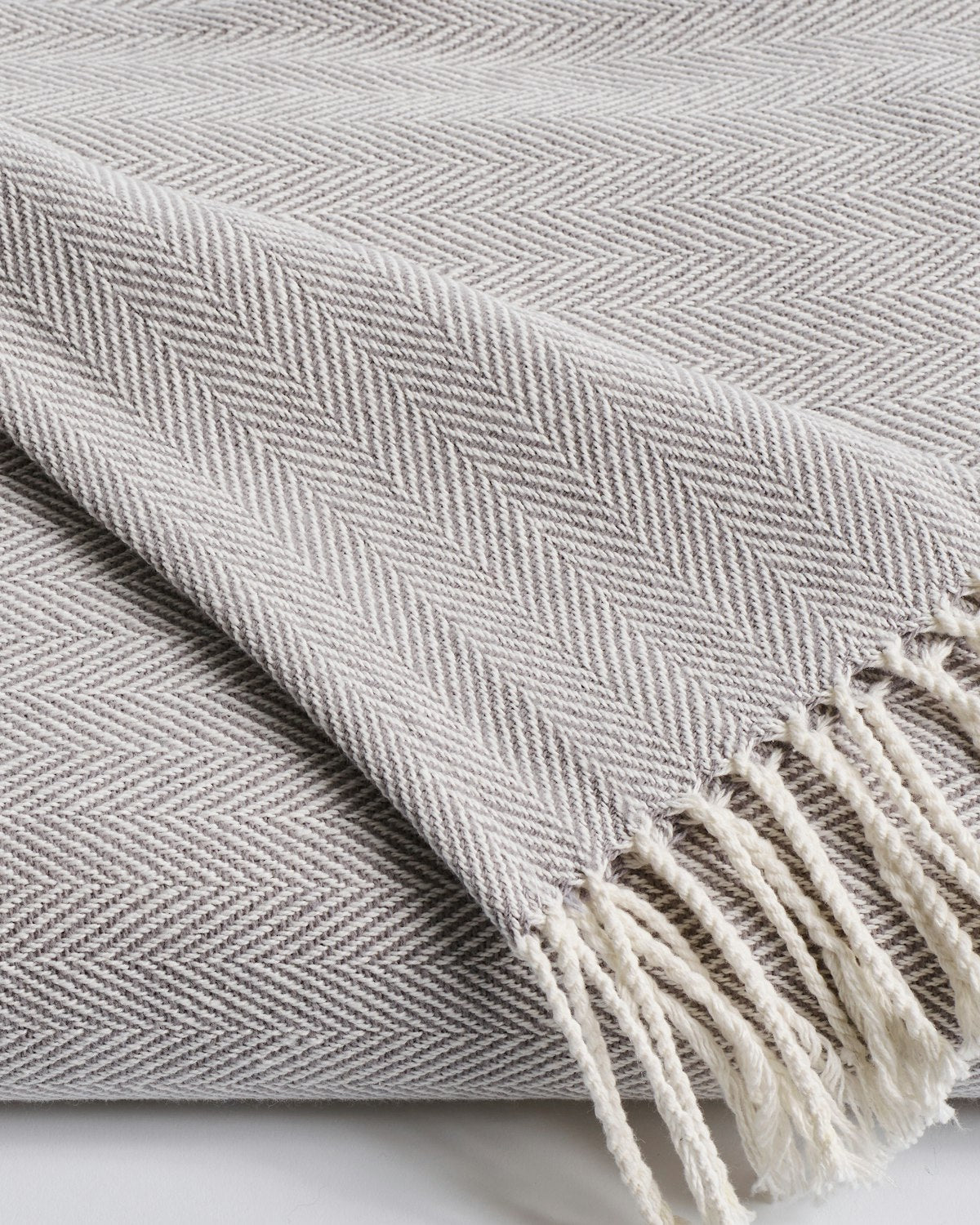 Close-up of a folded Herringbone Throw in Light Grey by Petite Plume, featuring an intricately woven zigzag design. The white fringe is neatly arranged, adding texture and contrast to the cotton fabric, making it a stylish addition for various home decor styles.