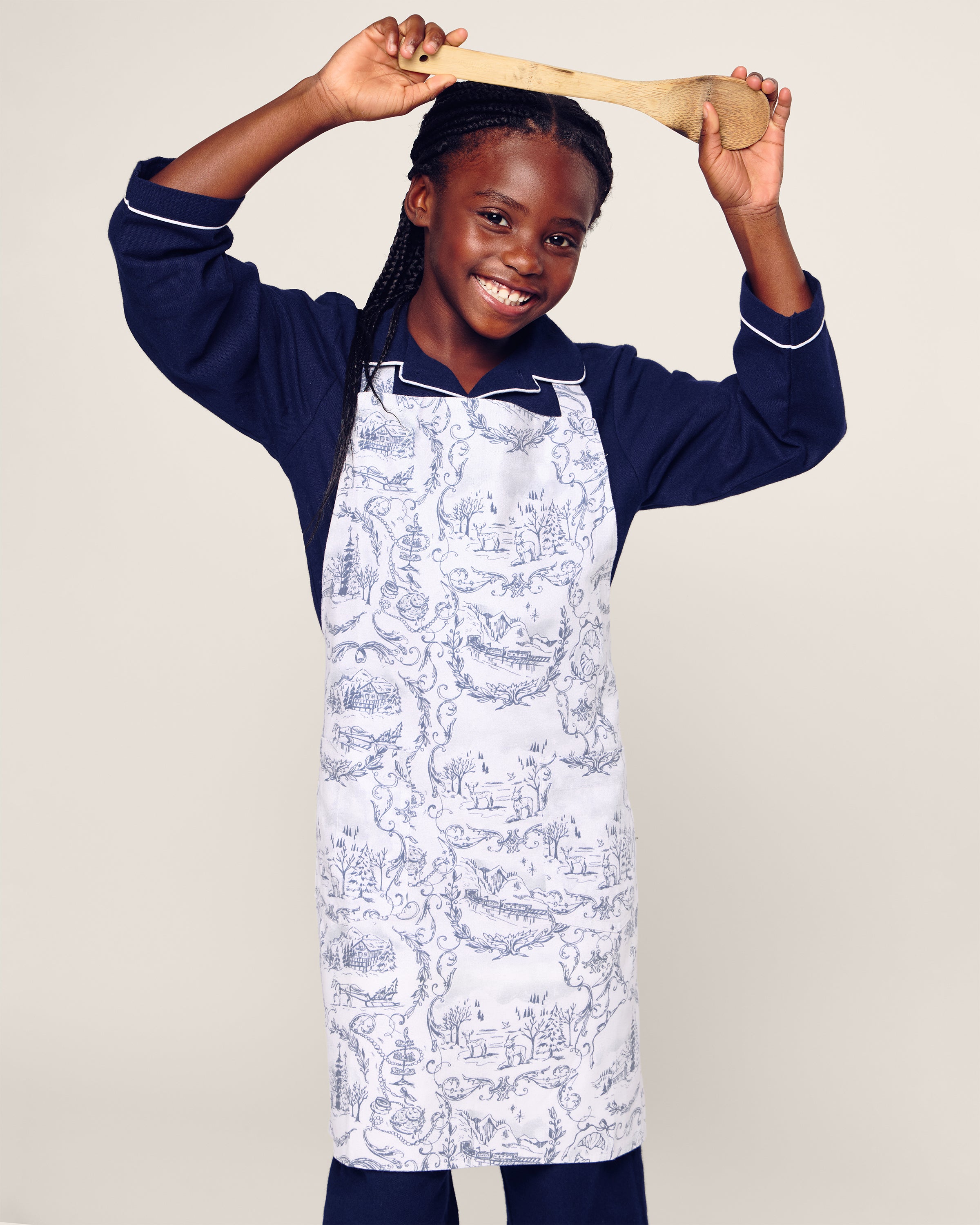 A cheerful child with braided hair wears a Maman x Petite Plume Alpine Toile Kids Apron over navy pajamas, raising a wooden spoon. This limited-edition apron by Petite Plume features intricate monochrome designs, perfect for holiday baking, set against a plain light background.