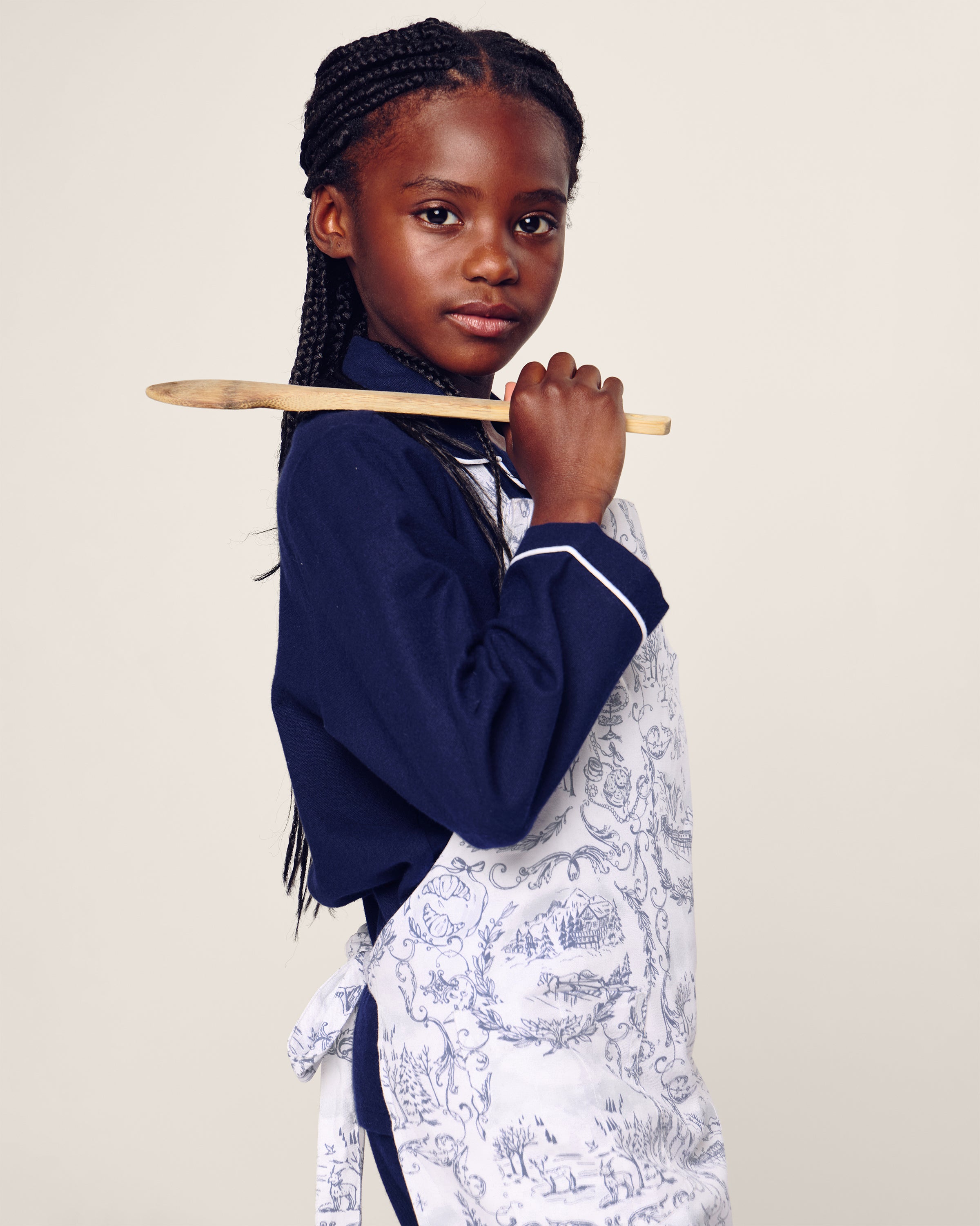 A young girl with braided hair in a dark shirt confidently poses, wearing the limited-edition Petite Plume Maman x Petite Plume Alpine Toile Kids Apron. She holds a wooden spoon over her shoulder against a neutral backdrop, ready for holiday baking.