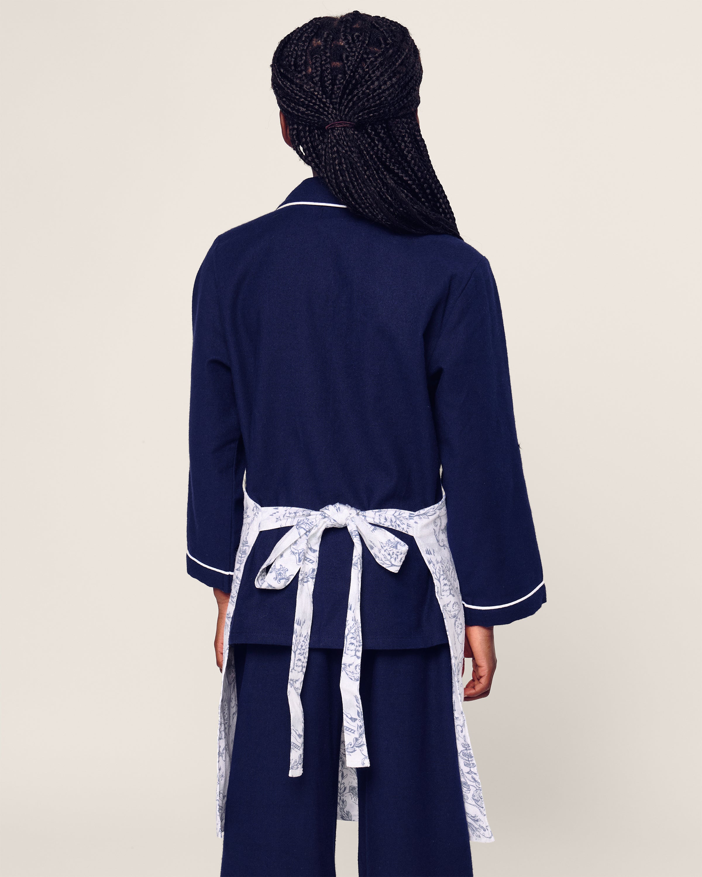 A person with braided hair, back turned, wearing a navy outfit with an open jacket and white accents featuring a limited-edition Petite Plume belt. The plain, light-colored background complements the style.