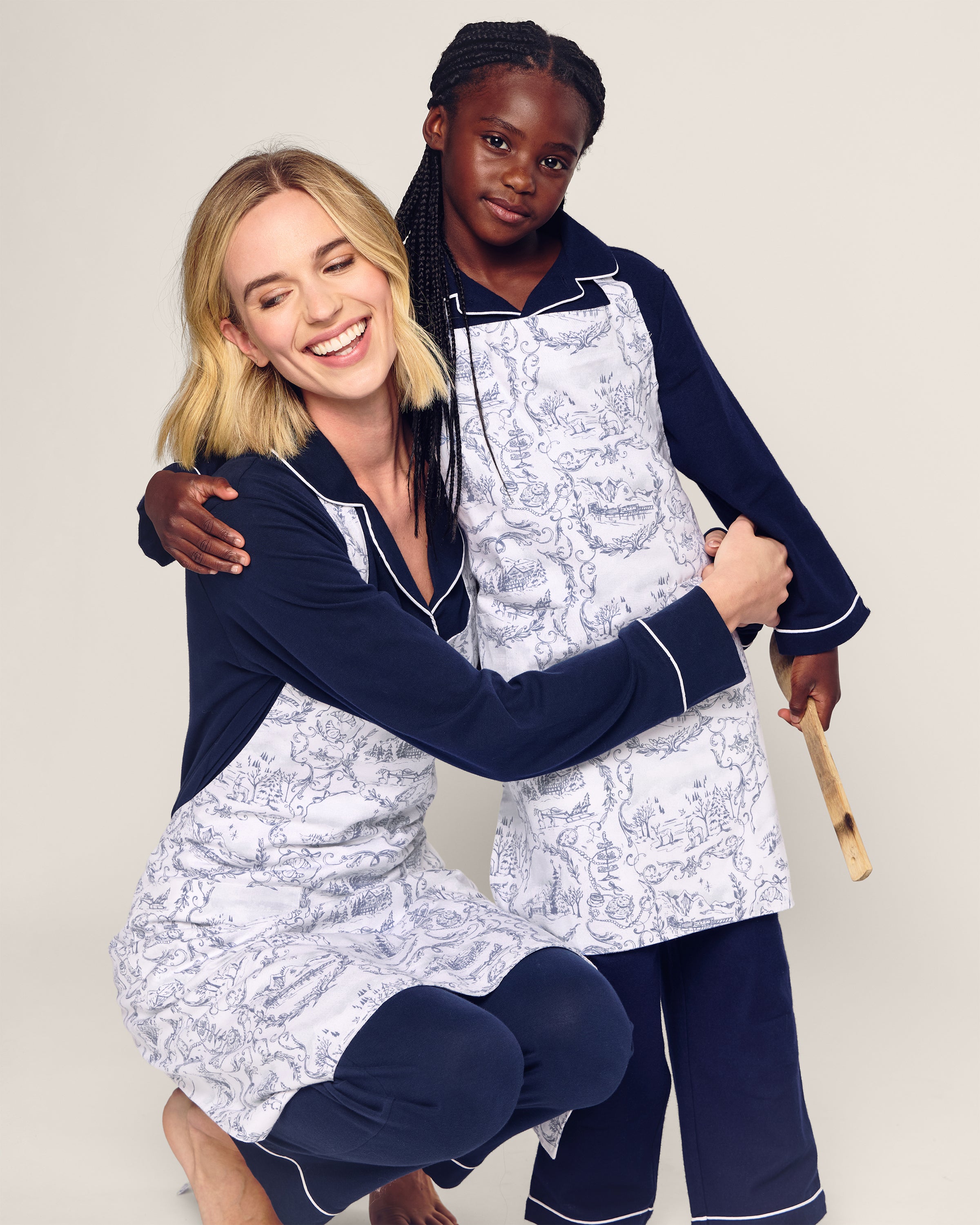 A woman and young girl smile and hug, wearing matching Petite Plume Maman x Petite Plume Alpine Toile Kids aprons and limited-edition blue pajamas. The woman kneels while the girl stands with a wooden spoon. Their aprons feature a white and blue floral design, perfect for holiday baking, against a plain background.