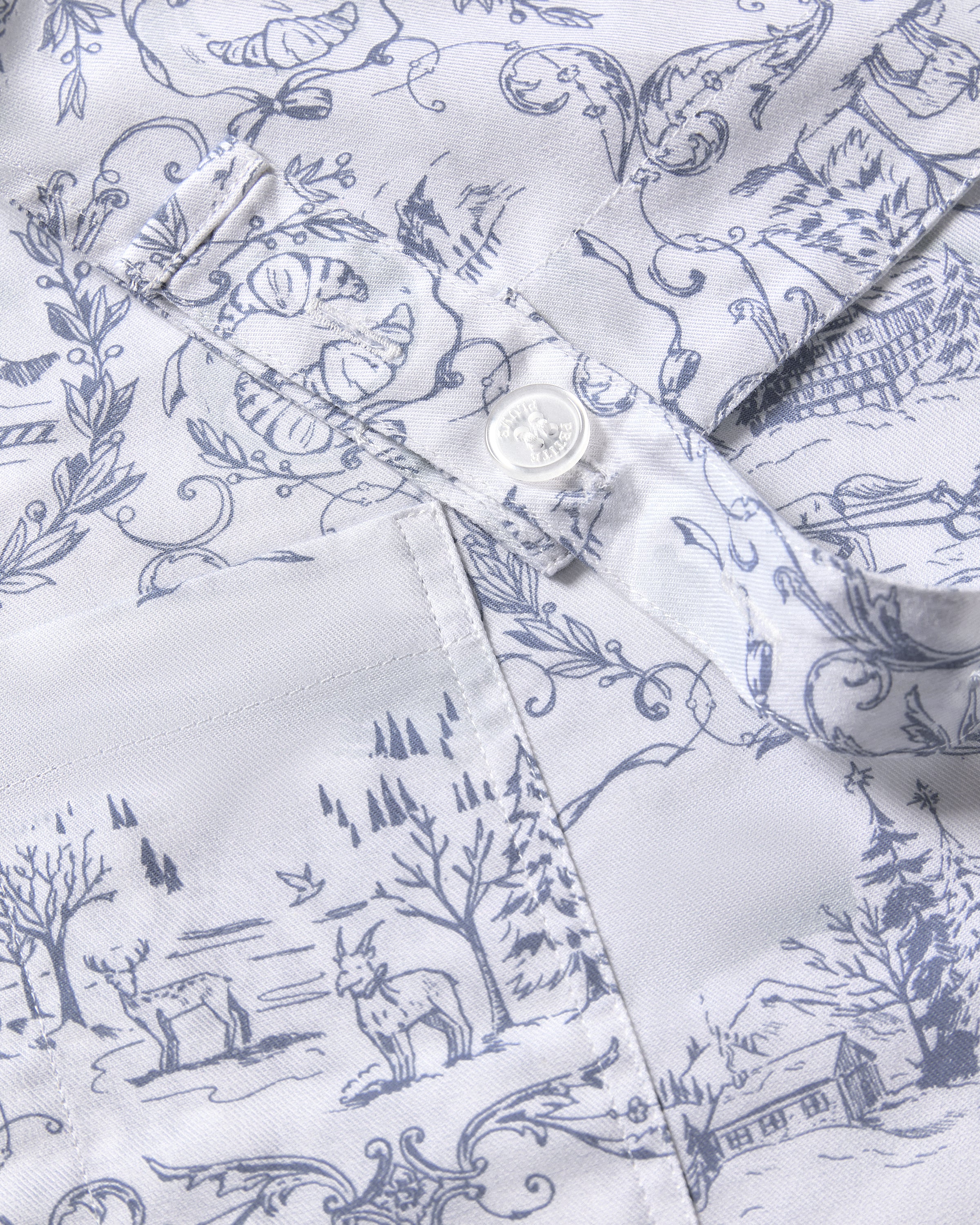 Close-up of the limited-edition Maman x Petite Plume Alpine Toile Kids Apron by Petite Plume, showcasing intricate blue designs with animals, trees, and countryside scenes on a white fabric.